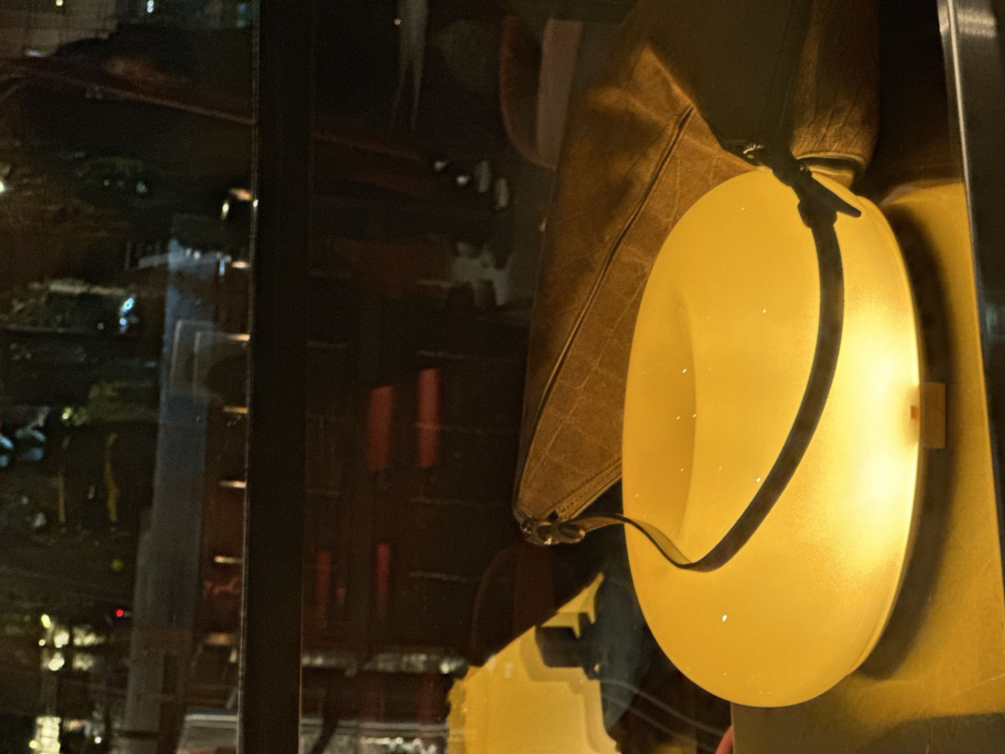 This image captures an indoor scene in Yerevan, Armenia, likely within a restaurant or cafe, looking out onto an urban environment at night. The foreground features a glowing, frosted yellow-orange, toroidal-shaped lamp with a dark, thin strap looping over its upper surface. The lamp rests on a reflective, dark-toned surface. Adjacent to the lamp on the right side, a brown, textured object, possibly a leather bag or cushion, is partially visible. The background, viewed through a window, presents a blurred urban nightscape with numerous scattered lights, indistinct silhouettes of trees, and dark building structures. Reflections on the window's surface superimpose vague interior elements, such as yellow shapes (possibly chairs) and dark furnishings, onto the exterior view. No individuals, specific actions, or interactions are clearly depicted.