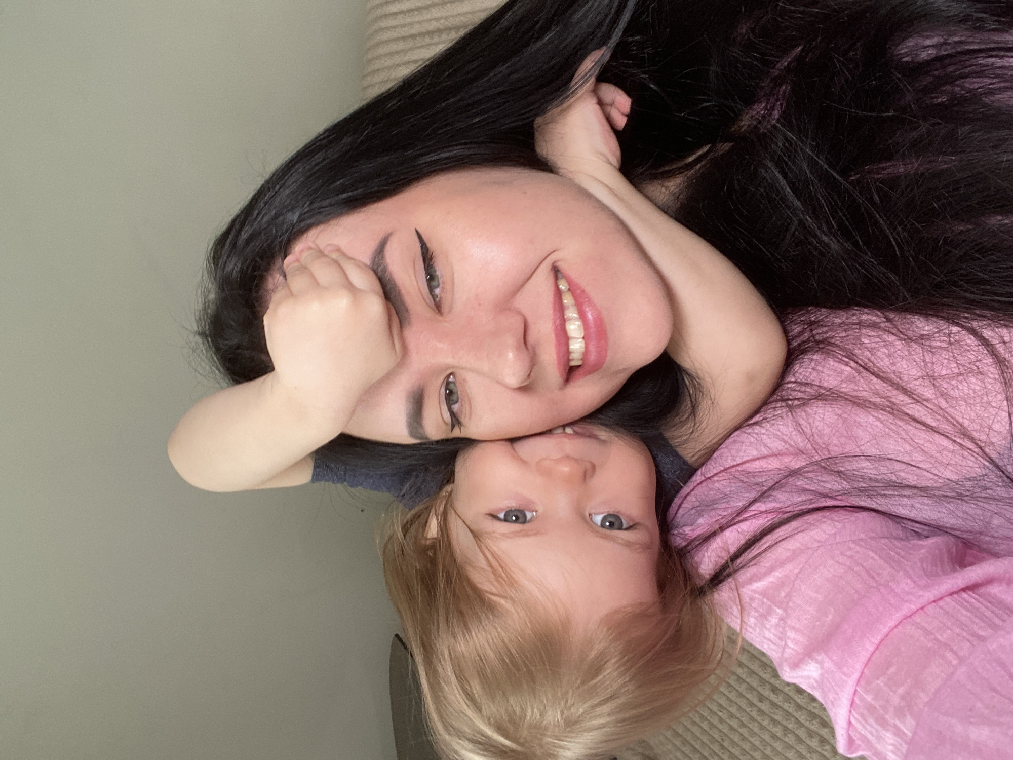 The image shows a woman and a child lying down together. The woman has long, dark hair with purple streaks and is smiling. She is wearing eyeliner. The child, likely a toddler, has light blonde hair and blue eyes, and is also smiling. Both are looking towards the camera. The background consists of a neutral-colored wall and what appears to be a textured surface, potentially a blanket or upholstery. The location cannot be identified as Chaikovsky, Russia, based solely on this image.