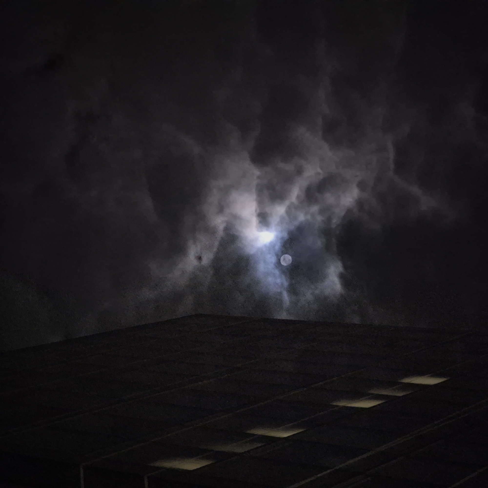 A low-angle, night-time photograph shows the moon partially obscured by clouds above a building. The moon is visible as a small, bright disk, surrounded by diffused light from behind the clouds. The clouds are dark and appear to be moving or swirling. The building is dark, with a flat roof visible. There are a series of light-colored rectangles, potentially skylights, that are spaced evenly across the roof. The location of the image is undetermined, but the city name is Strasbourg, France.
