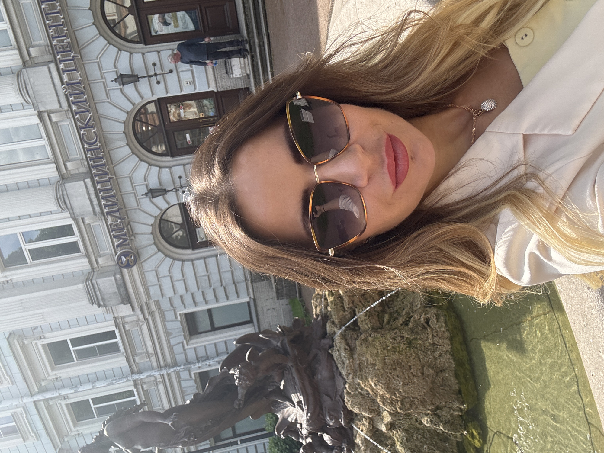 A woman with blonde hair, wearing sunglasses and a light-colored blazer, takes a selfie in front of a building. A bronze statue of a mythical creature and a fountain are to her left. The building has a sign that reads "МЕДИЦИНСКИЙ ЦЕНТР" (Medical Center) in blue lettering and ornate architectural details. A man in a dark shirt and pants stands on the steps of the building in the background. The scene appears to be in Saint Petersburg, Russia, likely near a medical facility.