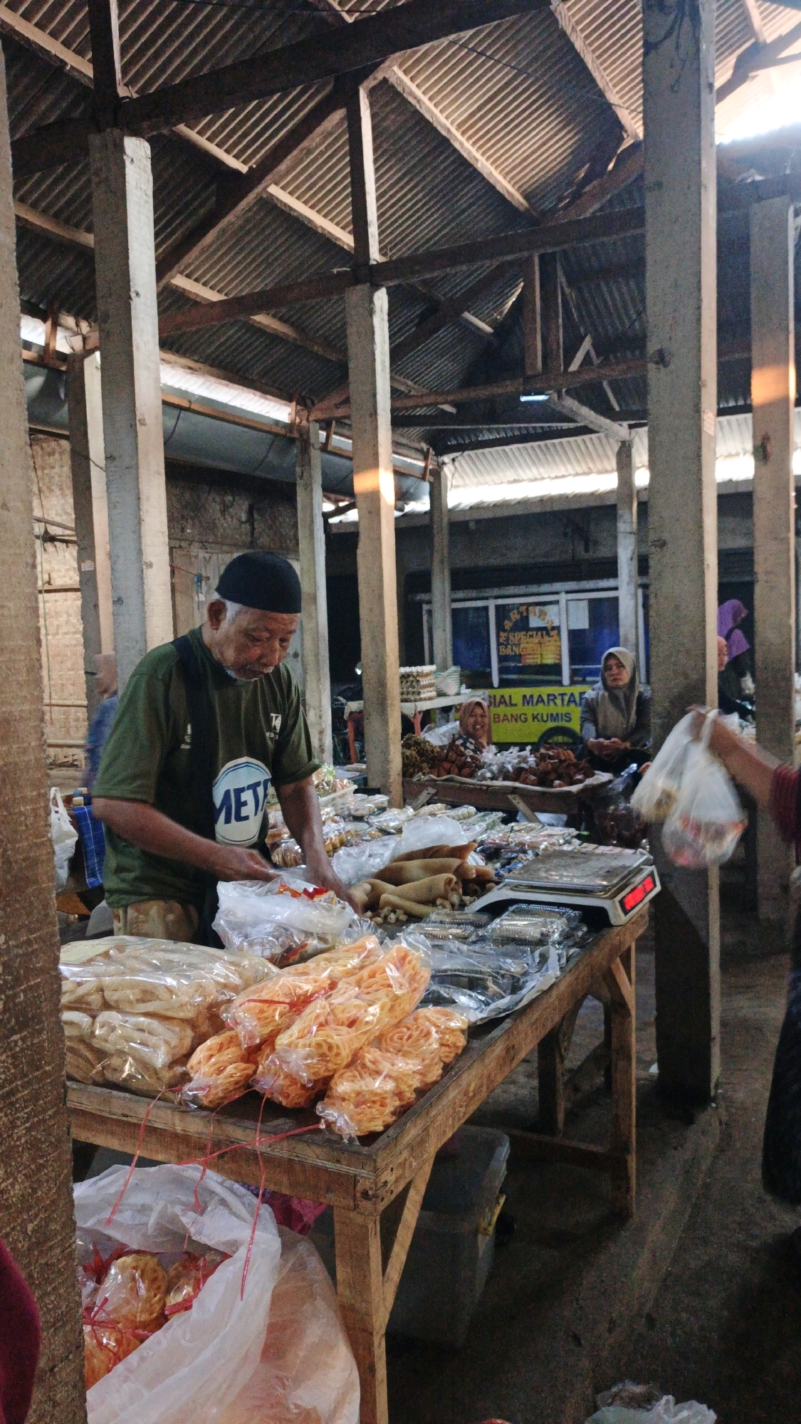 A man wearing a black skullcap and a green t-shirt with "META" printed on it is standing behind a wooden stall in an open-air market in Cibenda, Indonesia. He appears to be a vendor, as he is handling plastic bags filled with food items. The stall is laden with various packaged goods, including what appear to be snacks like crackers and fried items, as well as some dried goods. Behind him, other vendors are visible, including a woman in a headscarf smiling, and a stall with eggs in crates. The market structure consists of a corrugated metal roof supported by concrete pillars. The ground is a concrete floor. The lighting suggests it is daytime.