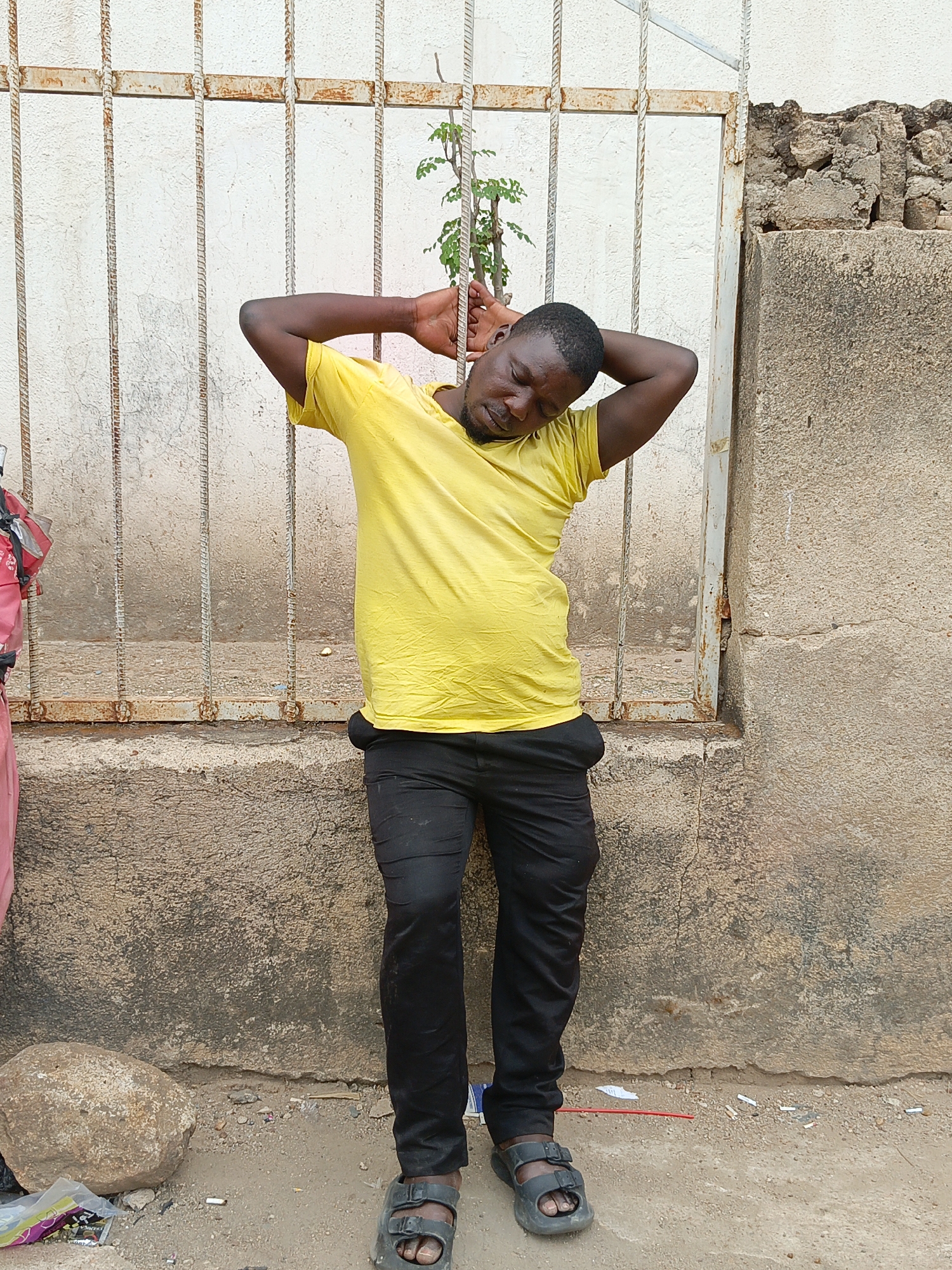The image shows a dark-skinned man standing with his back against a rusty, metal-barred gate in what appears to be Jos, Nigeria. He is wearing a yellow t-shirt, black pants, and black sandals. His hands are behind his head, and he is looking down with his eyes closed. A small green plant is growing through the bars of the gate behind his head.
The gate is set into a weathered concrete wall, which is cracked and stained. To the left of the gate, part of a person in pink is visible. The ground is a mixture of dirt and gravel, with scattered trash including cigarette butts and bits of plastic. A large rock is to the left. The wall on the right is made of concrete blocks.