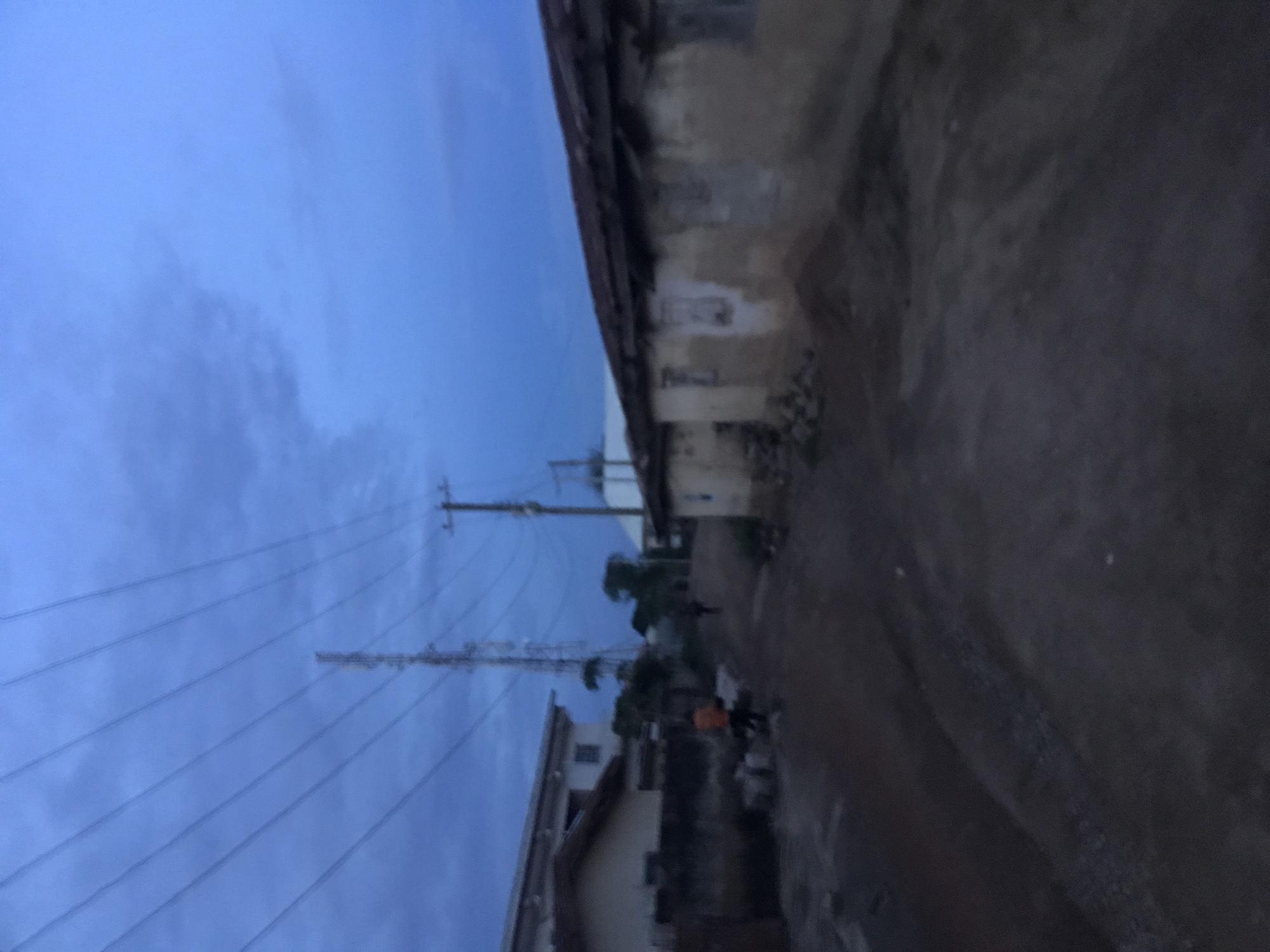This image depicts a street scene in Rafin Par, Nigeria, under low light conditions consistent with dawn or dusk, characterized by a deep blue sky with scattered clouds. The foreground and midground are dominated by an unpaved, dark brown dirt road that recedes into the distance.
On the left side of the road, a two-story building with a white facade and a brown roof is partially visible, behind a lower single-story structure featuring a lighter facade and a dark roof. A dark wall or fence runs along the roadside in front of these buildings. A pile of light-colored construction blocks is present on the ground. An individual, identifiable by an orange upper garment, stands near these blocks, facing away from the viewer. Further down the road, another person is seen walking away.
The right side of the road features an older, single-story building with weathered, light brown or grey mud-plastered walls and a dark, possibly corrugated metal, roof. Piles of debris or stones are visible at the base of this structure. Darkened openings, likely windows, are present on its facade.
In the mid-distance, a tall metal lattice tower, consistent with a telecommunications or power transmission tower, stands against the sky. To the right of the lattice tower, a wooden utility pole with multiple crossarms supports numerous overhead wires that traverse the upper portion of the frame. The immediate environment around the road and structures appears to be unkempt, with varying textures of dirt and debris.