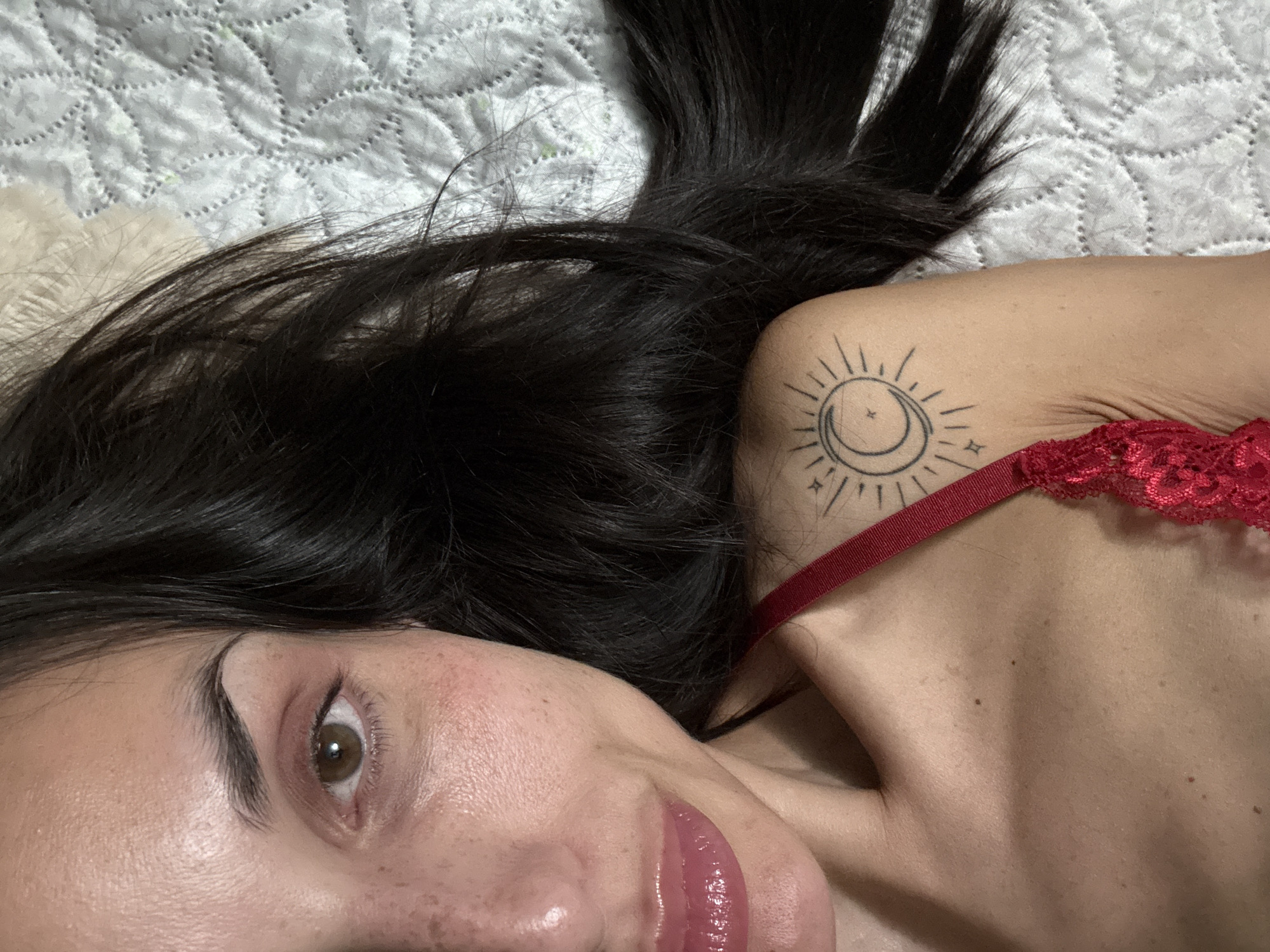 The image shows a woman lying on a quilted surface. She has dark hair and light skin. A tattoo of a sun and moon is visible on her shoulder. She is wearing a red lace garment. Her face is close to the camera, with focus on her eye and lips. There is no information to determine the location is in Pleven, Bulgaria.