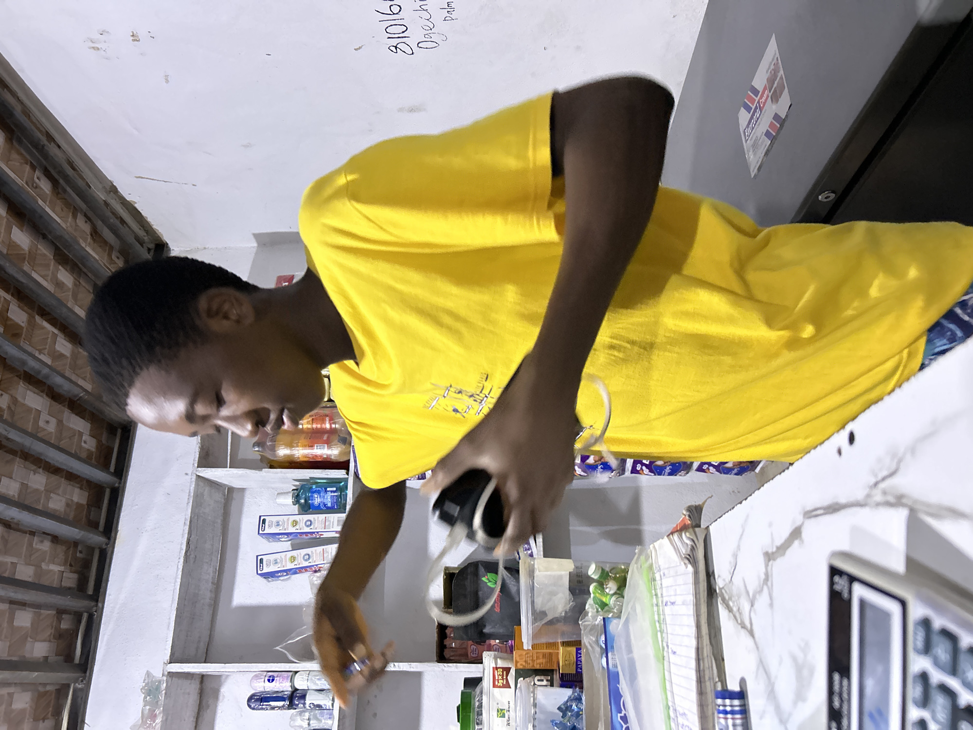 An individual in a yellow t-shirt is holding a charging adapter while standing behind a counter filled with various items. The items on the counter appear to be goods for sale, including small packaged items. Behind the individual, a shelf is stocked with various household and personal care products. The location is identified as Ihiagwa, Nigeria.