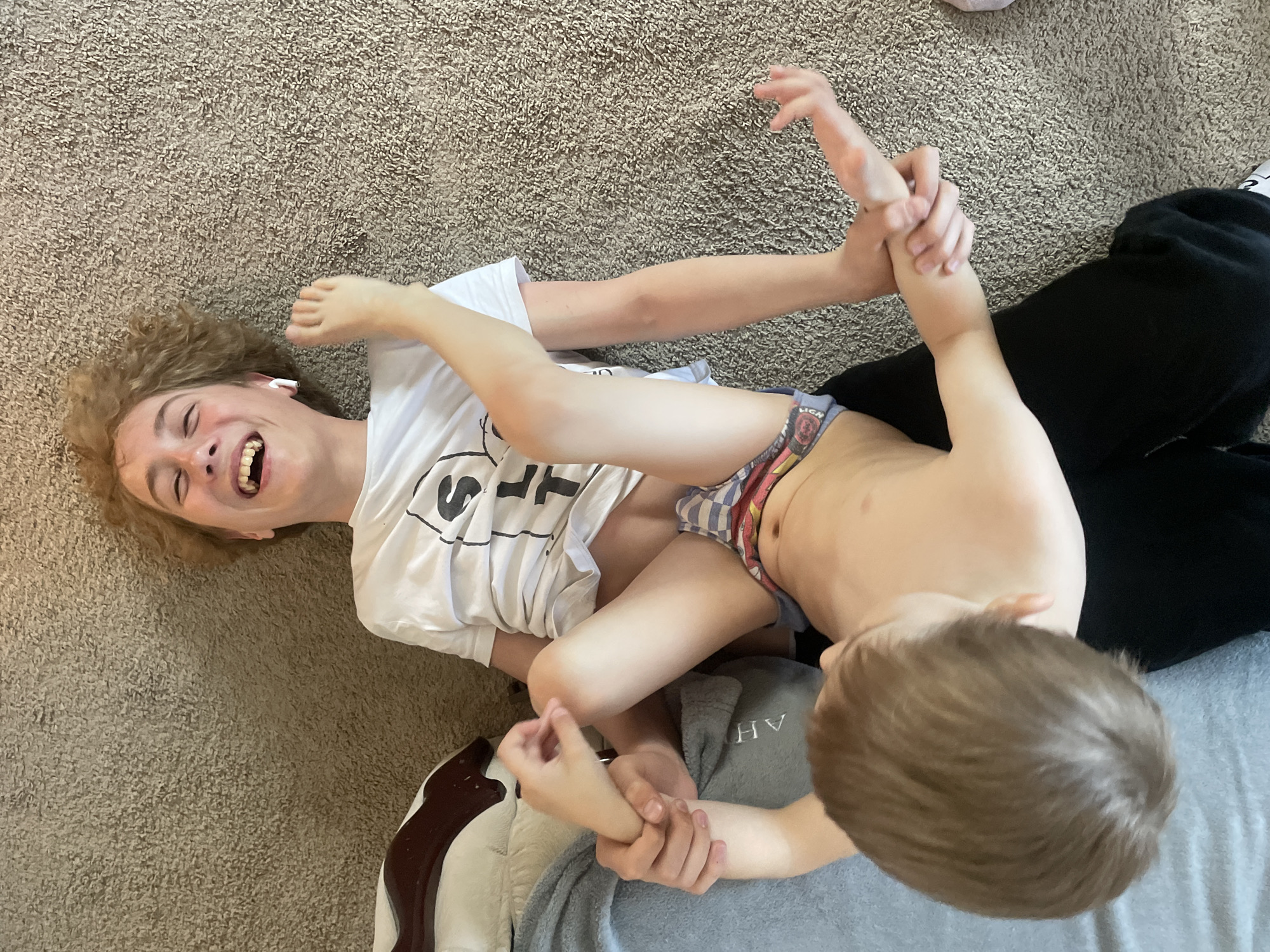 Two people are wrestling on a carpeted floor. One person is lying on their back, wearing a white t-shirt and earbuds, and laughing. The other person is partially undressed, wearing only underwear. Their limbs are intertwined, and they are gripping each other's arms and legs. There are blankets under them and next to them. The location is not identifiable.