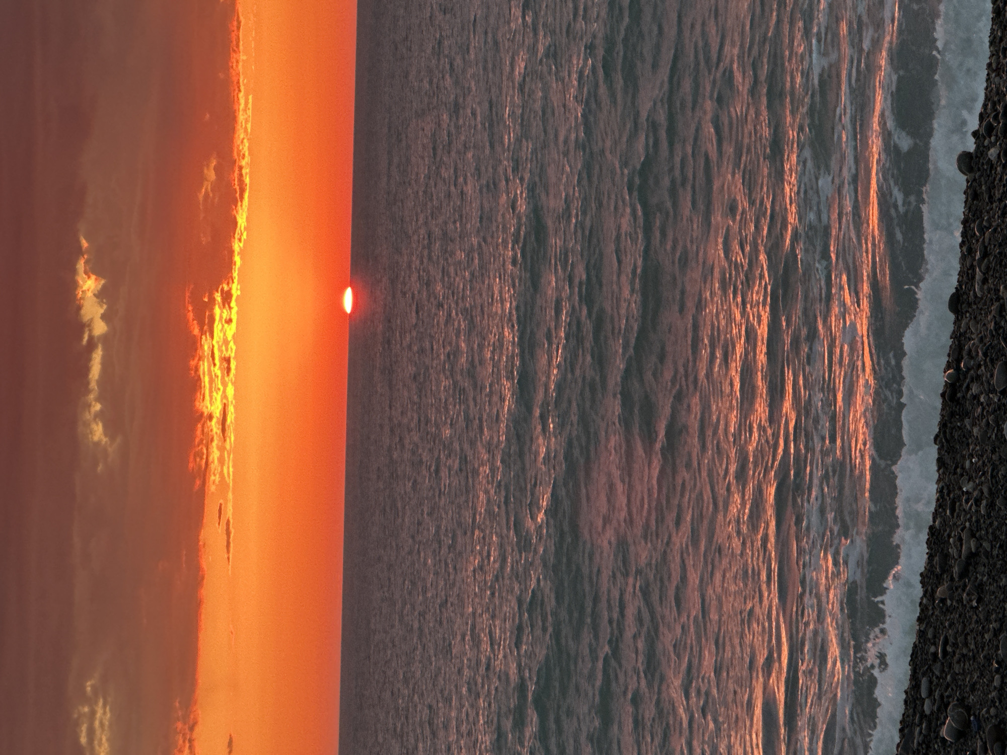 The image shows a sunset over the Black Sea, likely taken in Batumi, Georgia. The sky is orange with clouds illuminated by the setting sun. The sun itself is a small, bright red-orange disc. The sea reflects the colors of the sunset. The shoreline is visible on the right edge of the image and is composed of dark pebbles. Waves are breaking on the shore.
