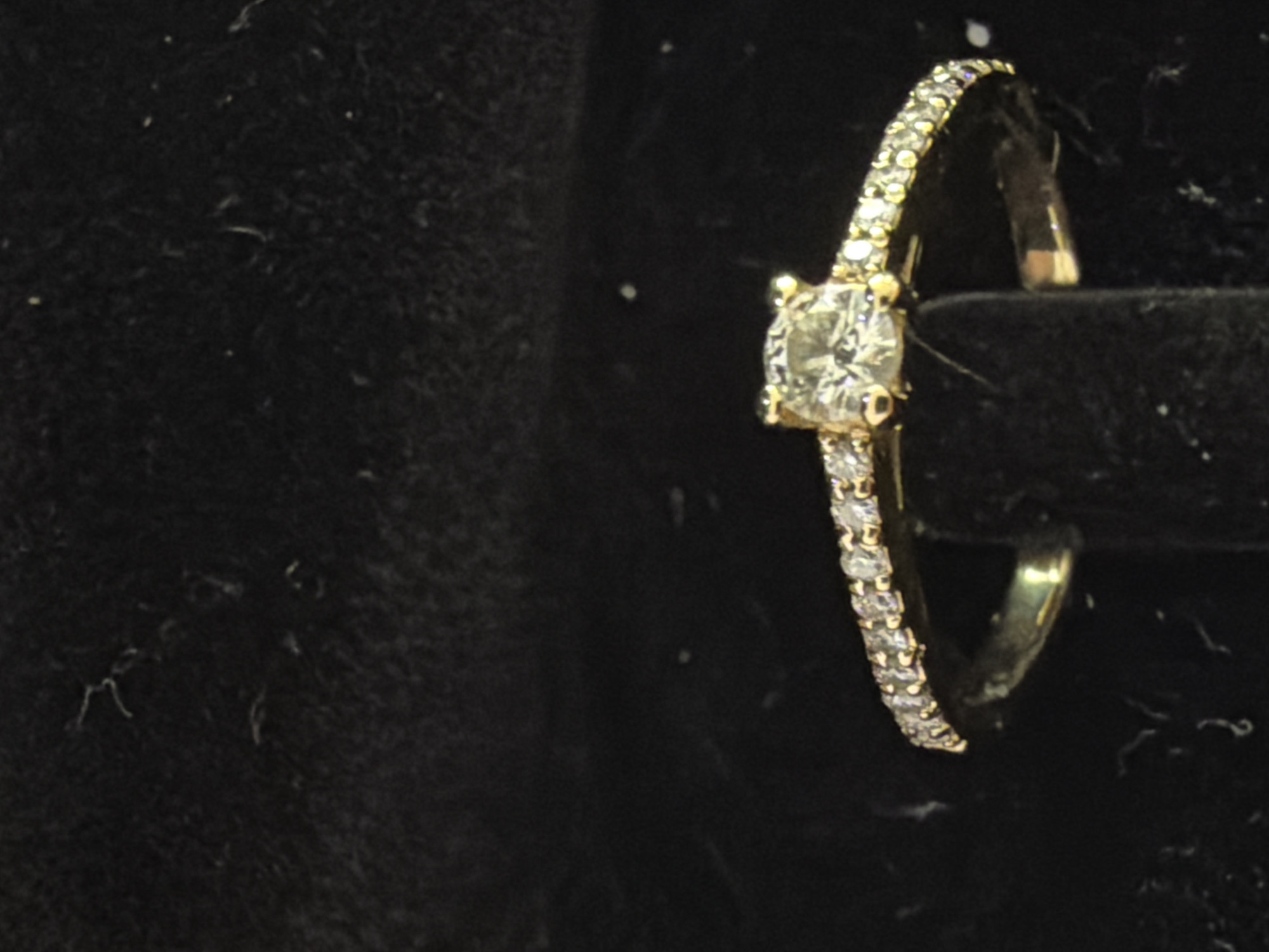 A gold ring with a central diamond is displayed on a black velvet background. The ring is encrusted with small diamonds along its band, and the central diamond is square-cut and appears to be a solitaire. The background is dark and slightly textured, with some lighter specks visible. The image is taken from a slightly elevated angle, focusing on the ring. There is no indication of a specific location in Sofia, Bulgaria.
