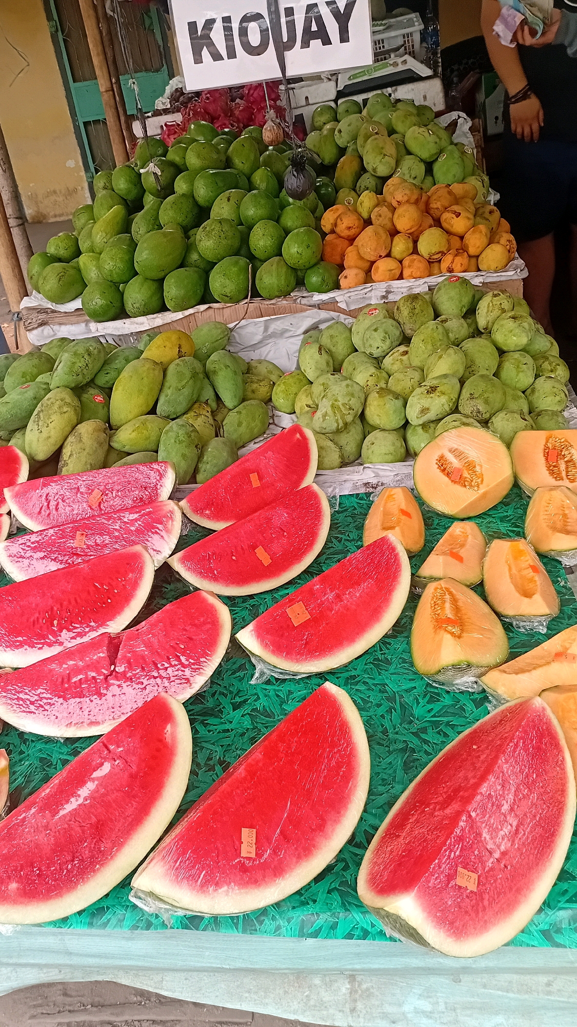 The image depicts an outdoor fruit stall in Kasang Kumpeh, Indonesia. A variety of fruits are displayed on tables covered with a green mat. In the foreground, several slices of watermelon are arranged, some wrapped in plastic. Behind the watermelons, there are wedges of cantaloupe, also wrapped in plastic. Further back, large piles of green mangoes and smaller piles of orange-colored fruits, possibly apricots or a variety of mango, are visible. In the background, red dragon fruit can be seen, along with a sign that reads "KIOJAY" in large black letters. A person's arm and hand, holding some papers, are visible on the right side of the image. The stall appears to be situated along a street, with wooden poles and a building visible in the background. The overall scene conveys a lively market atmosphere with an abundance of fresh produce.