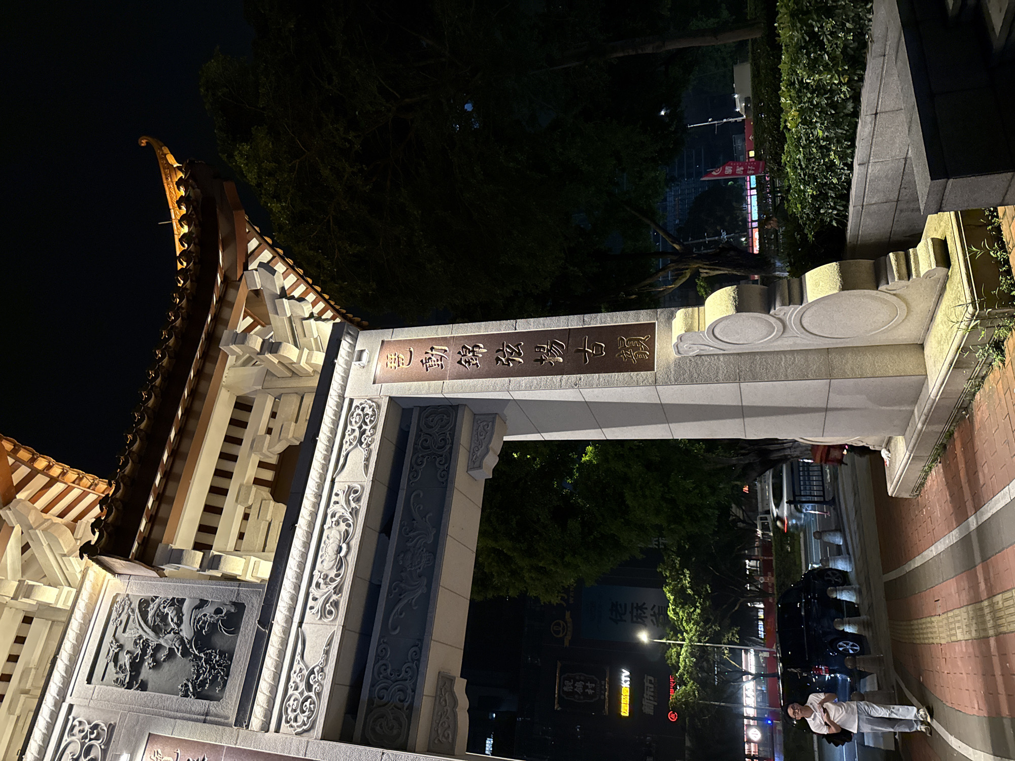 This media file captures a nighttime scene in Haizhu District, China, likely a street or a plaza area with traditional architectural elements. The dominant subjects are a large, ornately decorated archway and a person standing on a sidewalk.
The archway features intricate carvings and traditional Chinese architectural styling, with a curved, tiled roof illuminated by warm lighting. The stone pillars of the archway are adorned with detailed bas-relief sculptures and decorative patterns. Chinese characters are prominently displayed on the archway, including "鶴村" and "琶動錦發揚古寶," which translates to something like "Crane Village" and "Harmonious movement, brilliant development, promoting ancient treasures." Other visible text includes "WAN" and what appears to be business signage such as "【左照合KTV" and "新东方," suggesting a commercial area.
A person, dressed in casual attire (a white t-shirt and light-colored pants), is standing on a brick-patterned sidewalk to the right of the archway, facing away from the camera. They appear to be posing or resting, with their arms behind their back. The presence of parked cars and streetlights indicates an urban environment.
The time of day is clearly night, with dark shadows and artificial lighting creating a distinct atmosphere. The weather appears clear, allowing for good visibility of the illuminated structures and signage. The overall impression is one of a vibrant urban space that blends traditional aesthetics with modern life, particularly evident in the juxtaposition of the ancient-style archway and contemporary signage.