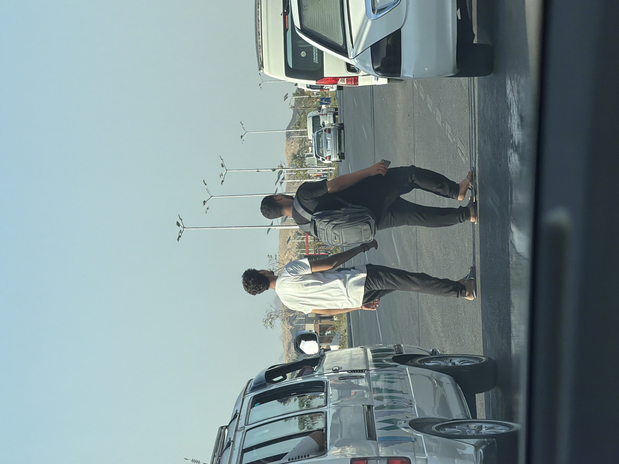 Two men are walking across a multi-lane road in Sharm El Sheikh, Egypt. The man on the left is wearing a white t-shirt and dark pants, and the man on the right is wearing a black t-shirt and black pants, with a grey backpack. They are walking in the same direction, away from the camera. A silver minivan is to the left of the frame, and a white sedan is to the right. The road is paved, and there are streetlights and some vegetation in the background. The sky is clear and blue.