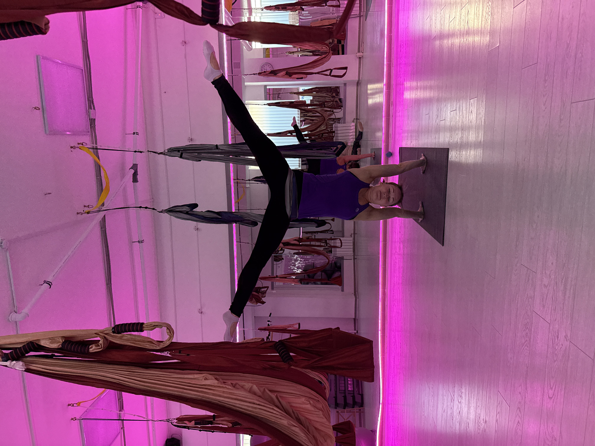 A woman is performing yoga in a studio lit with pink light. She wears black leggings and a purple tank top. Her legs are split into a wide V-shape, with one leg extended upward and the other downward. She is balancing on a yoga mat with her arms extended, one on each side of her body. The studio features aerial yoga equipment hanging from the ceiling. The room is illuminated by bright pink light, which is reflected off the wooden floor. Based on the location metadata provided in the request prompt, the location is Chaikovsky, Russia.