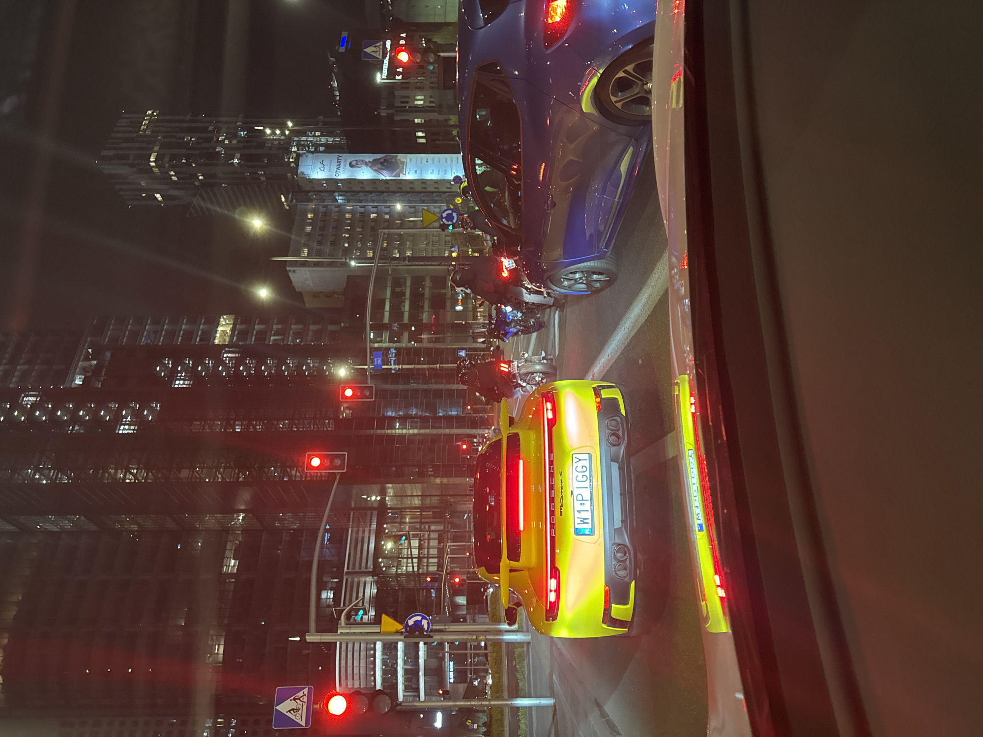 The image shows a nighttime urban scene in Warsaw, Poland. Cars and motorcycles are stopped at a red traffic light. The street is lined with modern high-rise buildings, and billboards. A yellow Porsche Carrera S with license plate "W1 P1GGY" is visible in the foreground, along with a blue car partially in the upper right corner. There are multiple motorcycles in the middle of the street.