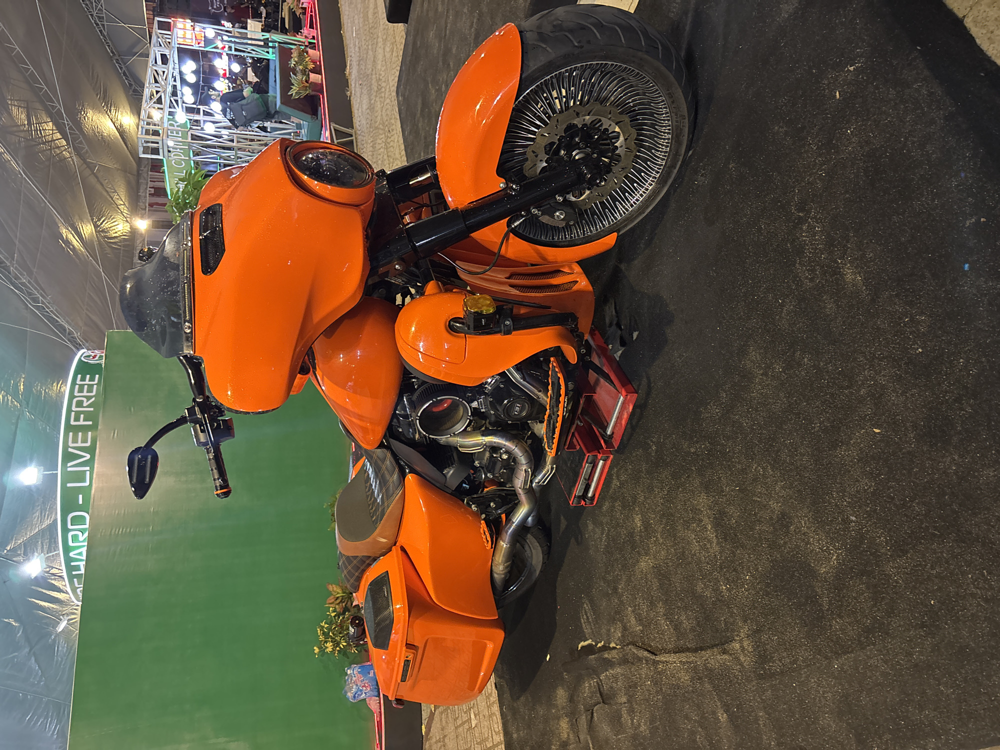 The image features a custom orange motorcycle prominently displayed within a structure that appears to be an exhibition hall. The hall's architecture includes a high, possibly tented roof, supported by visible framework. A green wall runs along the left side of the frame, marked with white lettering stating "RIDE HARD - LIVE FREE". In the background, partially obscured, is a multi-tiered display structure with bright lighting, potentially showcasing other products or exhibits. The floor surrounding the motorcycle is covered with a dark-colored material that appears textured. The overall setting suggests the scene takes place at an event or trade show featuring motorcycles, and that this display is located within Nha Trang Ward, Vietnam.