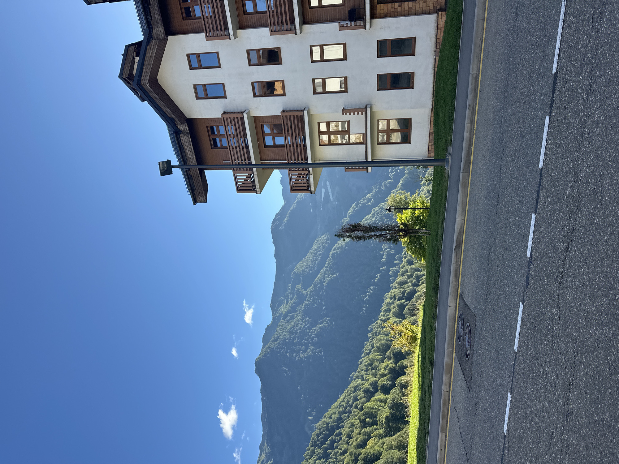 The image captures a building nestled against a backdrop of majestic mountains, likely in Estosadok, Russia. The building, with its white facade and wooden accents, features multiple stories with balconies and numerous windows. A streetlamp stands tall near the building. The scene is bathed in the clear, bright light of a sunny day, casting a serene mood.
The foreground is dominated by a paved road with distinct yellow and white markings, suggesting a well-maintained area. Beside the road, a strip of vibrant green grass separates it from the building. Beyond the manicured lawn, the imposing mountains rise, their slopes densely covered with lush green trees, indicating a forested natural environment. A few wispy white clouds dot the expansive blue sky, adding depth to the scenery. The overall impression is one of peaceful coexistence between human architecture and the grandeur of nature. The time of day appears to be late morning or early afternoon, given the quality of light and the absence of long shadows. There is no visible text in the image.