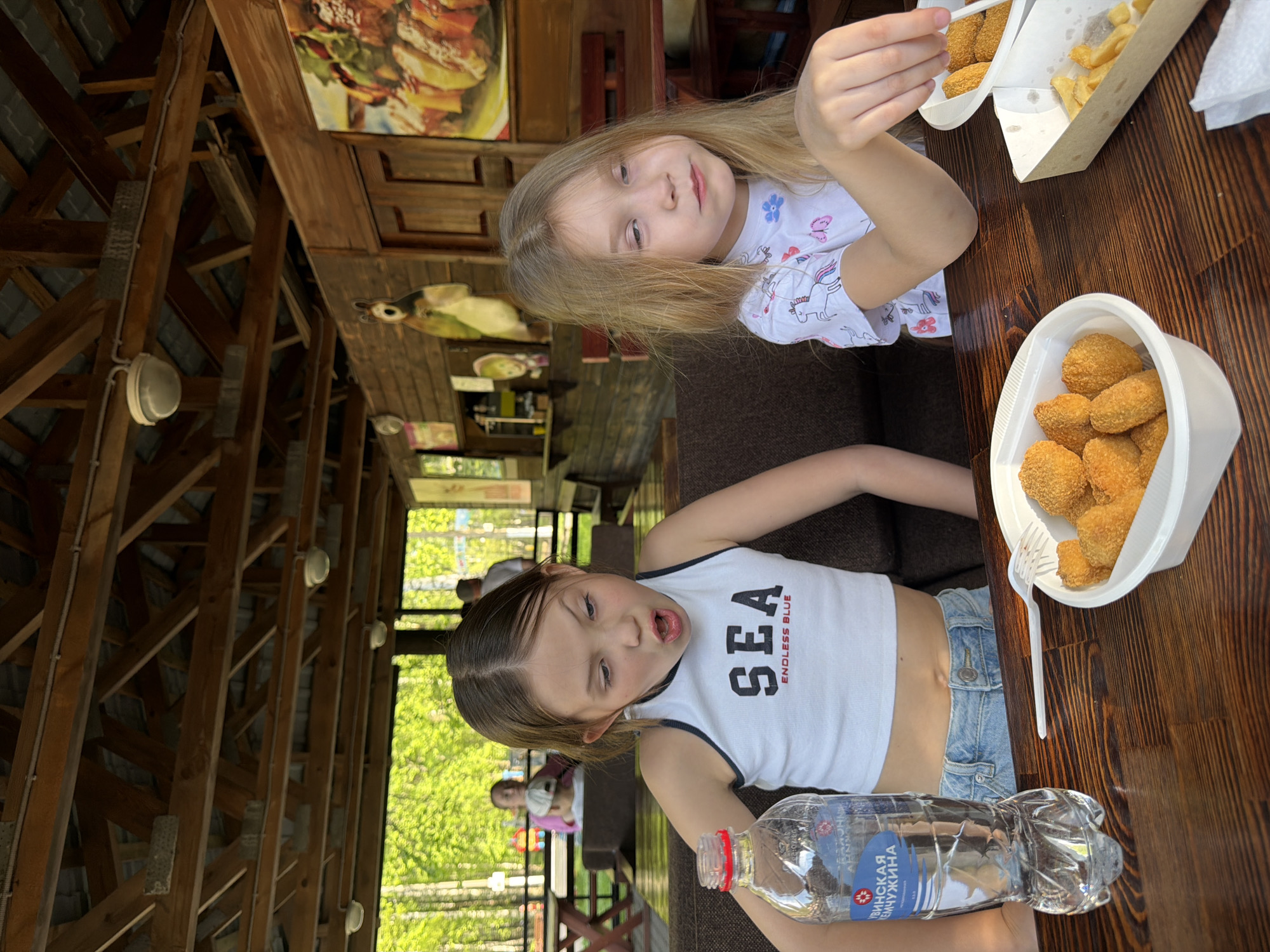Two young girls are sitting at a dark wooden table, presumably eating. The girl closest to the camera is wearing a white tank top with "SEA ENDLESS BLUE" printed on it and denim shorts; she has a plastic bottle of water near her. The second girl has blonde hair and is wearing a white T-shirt with a unicorn design. On the table are containers holding nuggets and fries. The scene appears to be taking place at an outdoor restaurant in Chaikovsky, Russia, as evidenced by the Cyrillic text on the water bottle. The structure of the open-air restaurant is visible, with wooden beams and a partially obscured view of trees in the background.