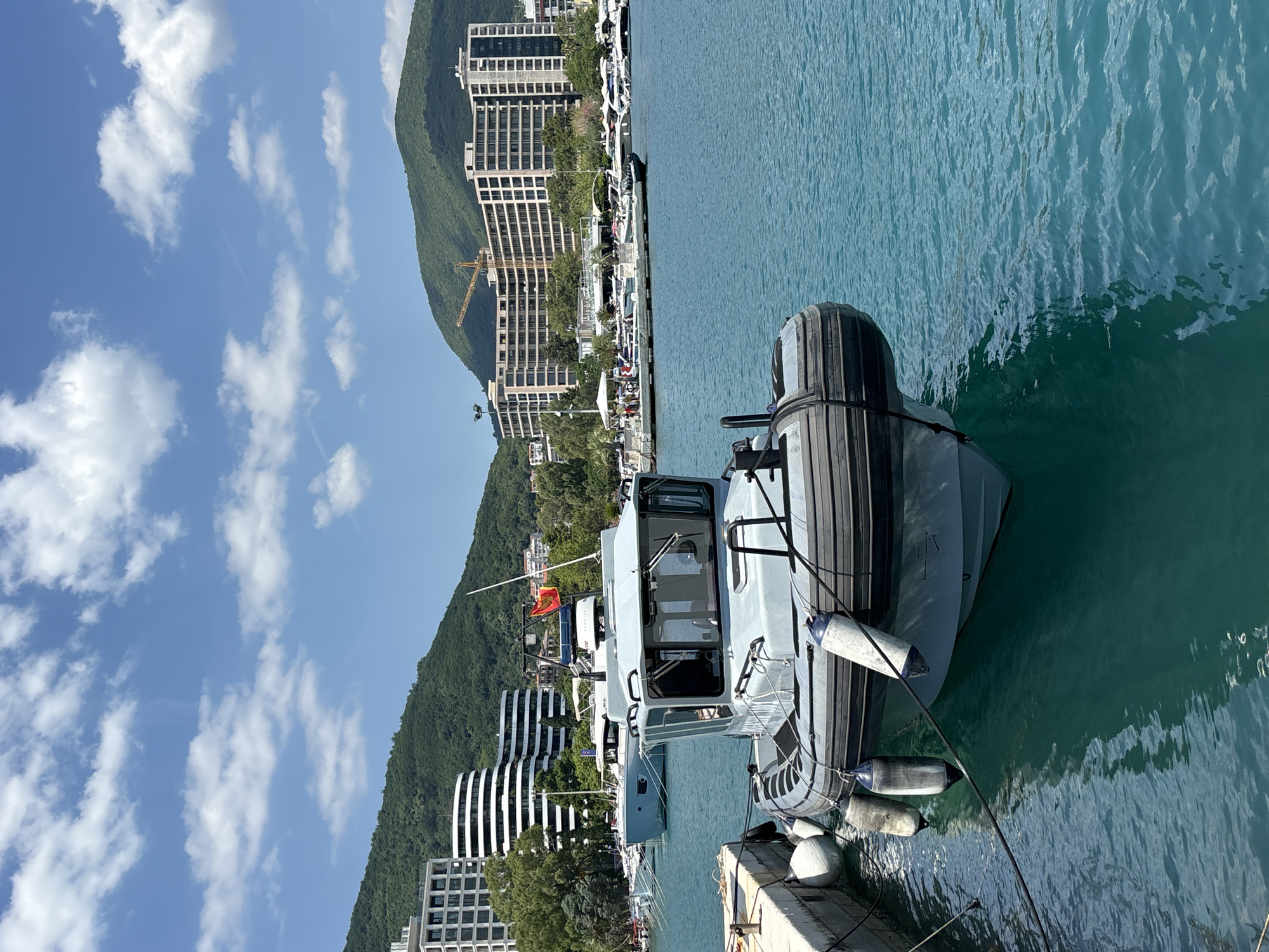 The image shows a marina in Budva, Montenegro, with several boats docked in the turquoise water. In the foreground, a rigid inflatable boat (RIB) is tied to a concrete pier. The RIB is light gray with a black inflatable collar and several white fenders hanging from its sides. A black rope connects the boat to the pier. In the background, a larger light blue yacht is docked behind the RIB. Further in the background, several other yachts and boats are visible. Behind the marina, modern multi-story buildings with curved architectural elements are built into a lush green hillside. A construction crane is visible among the buildings on the right side of the image. The sky is blue with scattered white clouds, and the sun is shining. The scene depicts a typical day at a coastal resort.