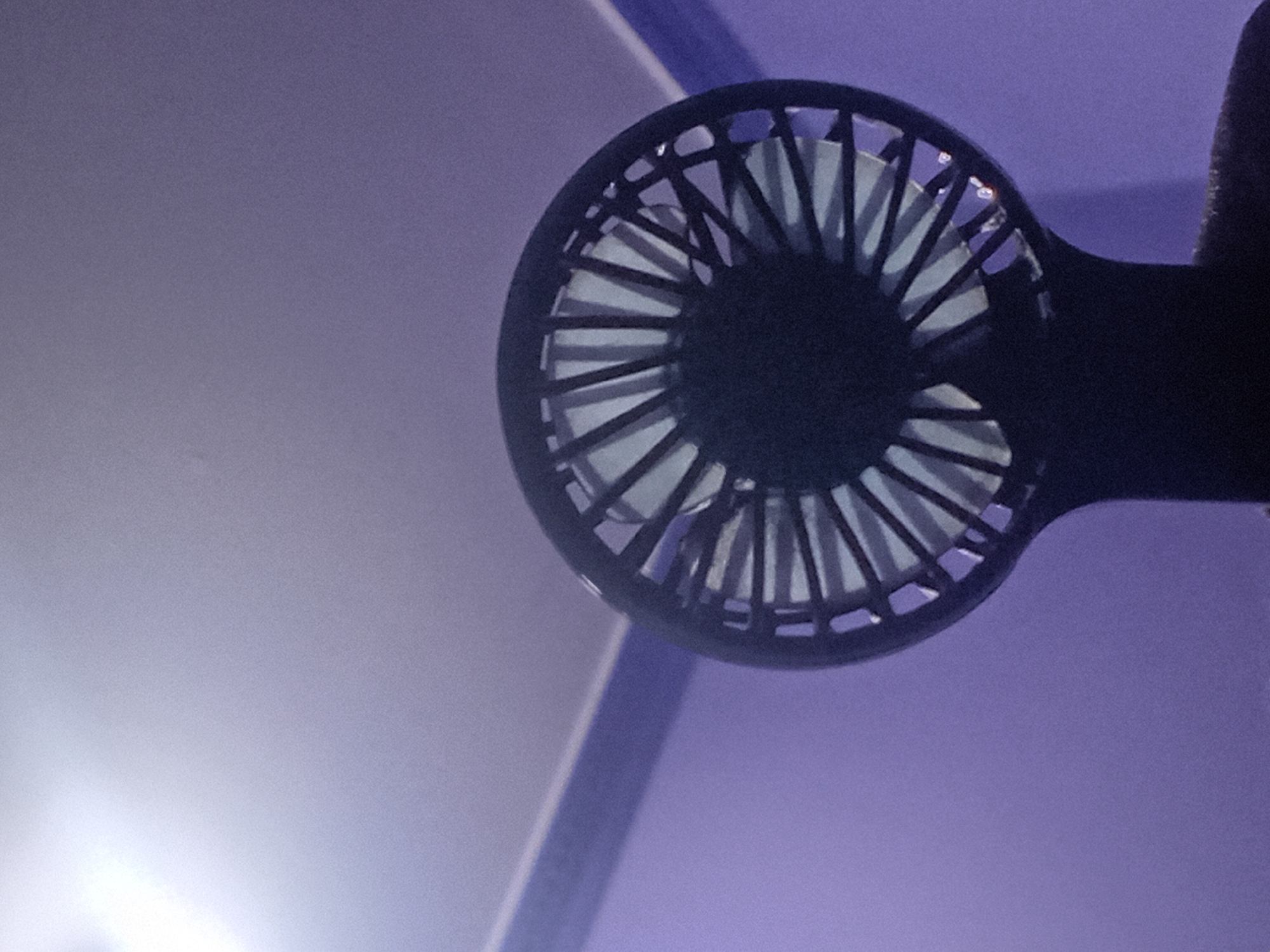 The image captures a close-up, low-angle shot of a black, portable fan. The fan's blades are visible through a black grille. The background consists of a light purple wall and a white ceiling, with a blue trim separating them. The lighting suggests an indoor setting, possibly a room. The fan appears to be positioned on a surface, indicated by a dark, textured material at the bottom of the frame. There are no people or discernible actions in the image. The scene is static and focuses solely on the object and its immediate surroundings. The location of this image in North Jakarta, Indonesia is not specified or inferable from the visual information provided.