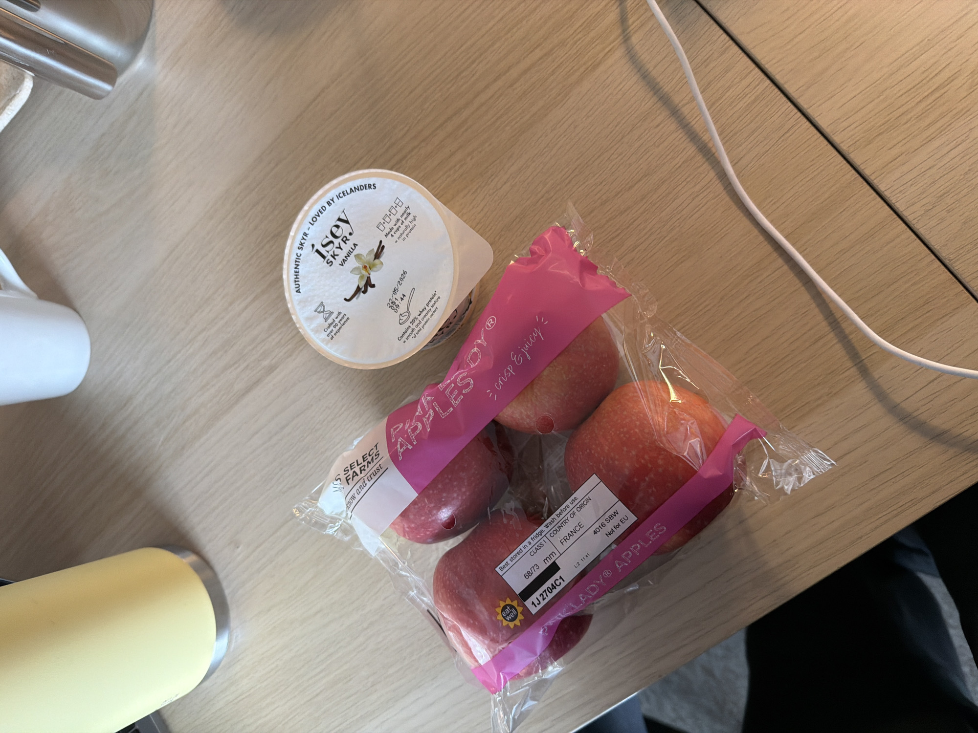 The image shows a table with an Isey Skyr Vanilla yogurt container and a package of Pink Lady apples. The yogurt is in the upper left of the frame and the apples are in the center. The packaging indicates the apples are from France. There are no people visible. The location is not identifiable as London, United Kingdom.