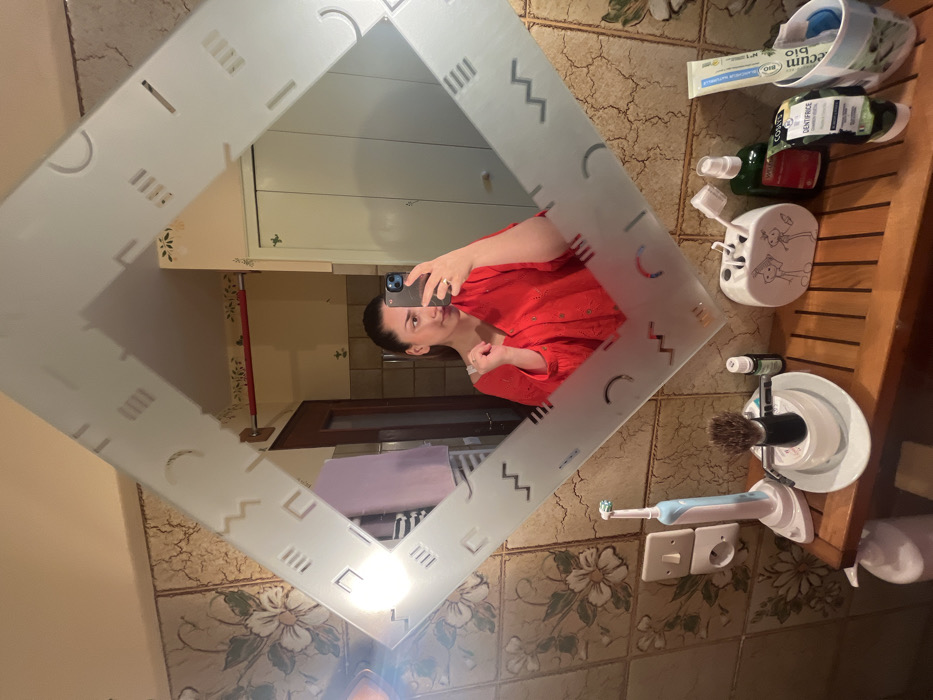The image captures a female individual taking a mirror selfie inside a bathroom located in Ancenis, France. The subject, visible from the chest up, is reflected in a diamond-shaped mirror framed with frosted geometric patterns. She is wearing a red, collared garment and holds a dark-colored smartphone in her right hand to capture the reflection. A ring is present on her right ring finger. Her dark hair is pulled back, and her left hand is positioned near her shoulder.
The reflected background behind the individual shows a light brown tiled wall with floral patterns on the left, a wooden door, and a white paneled door or cabinet on the right. A red horizontal bar is mounted on the wall. A light source on the far left creates a bright glare in the mirror.
The foreground displays a wooden slatted countertop holding various personal care items. From left to right: a light blue and white electric toothbrush on its charging base, plugged into a white double electrical outlet; a shaving brush with a dark handle in a white stand; a small dark bottle; a white ceramic toothbrush holder decorated with two cartoon figures, containing multiple toothbrushes; a green spray bottle; and a tube labeled "DENTIFRICE BIO" (organic toothpaste) in a white container. The wall directly behind these items is tiled with a textured, light brown surface.