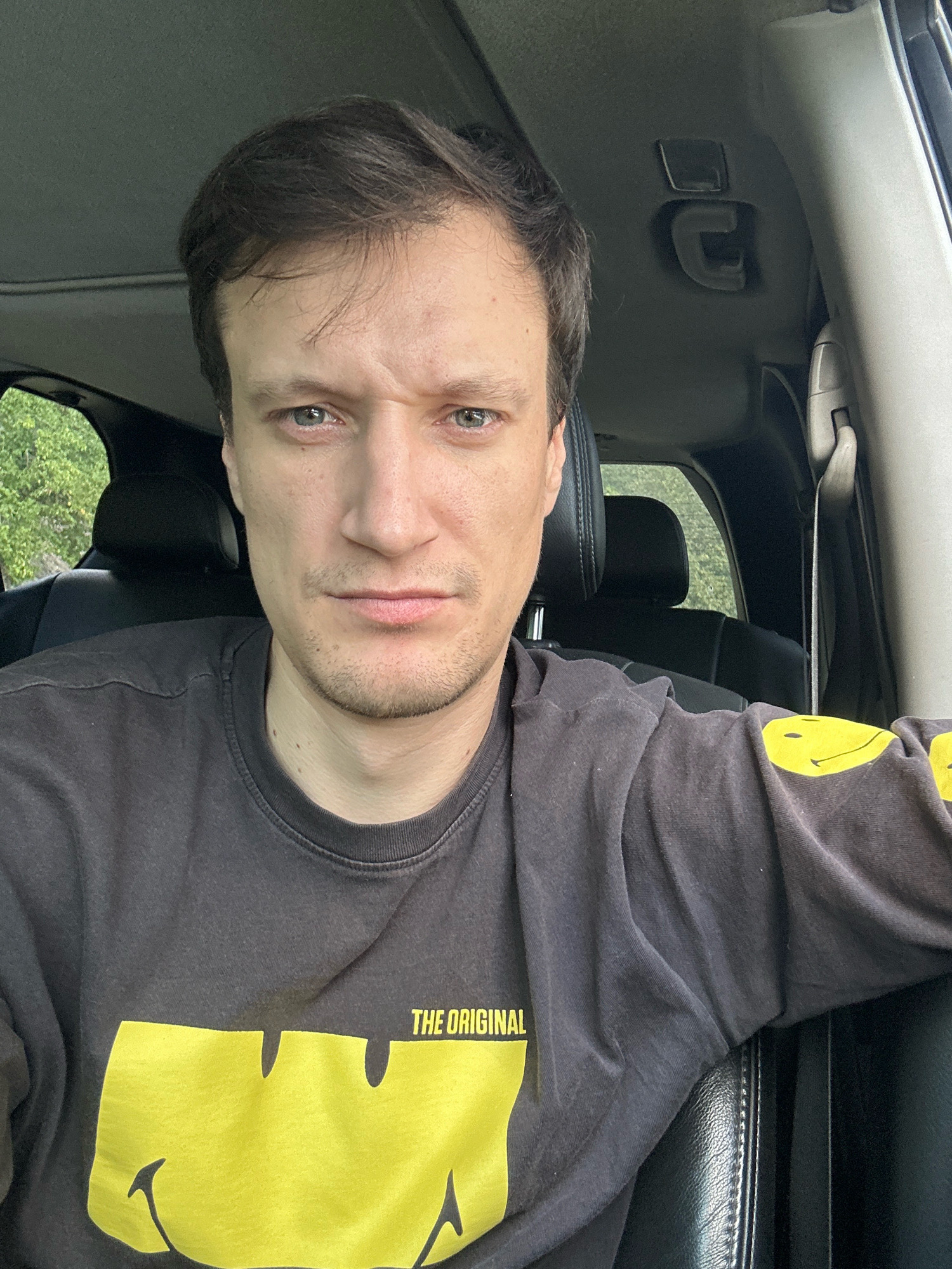 A man with short, dark brown hair, light blue eyes, and a neutral expression sits in the driver's seat of a vehicle. He is wearing a dark gray long-sleeved shirt with a large yellow smiley face graphic on the front and a smaller yellow text graphic that reads "THE ORIGINAL" above it. The interior of the vehicle appears to be black leather. Outside the vehicle, lush green trees are visible through the windows. The image is a selfie, taken from the man's perspective. No specific location in the city of Makiv, Ukraine, can be identified from the visual information provided.