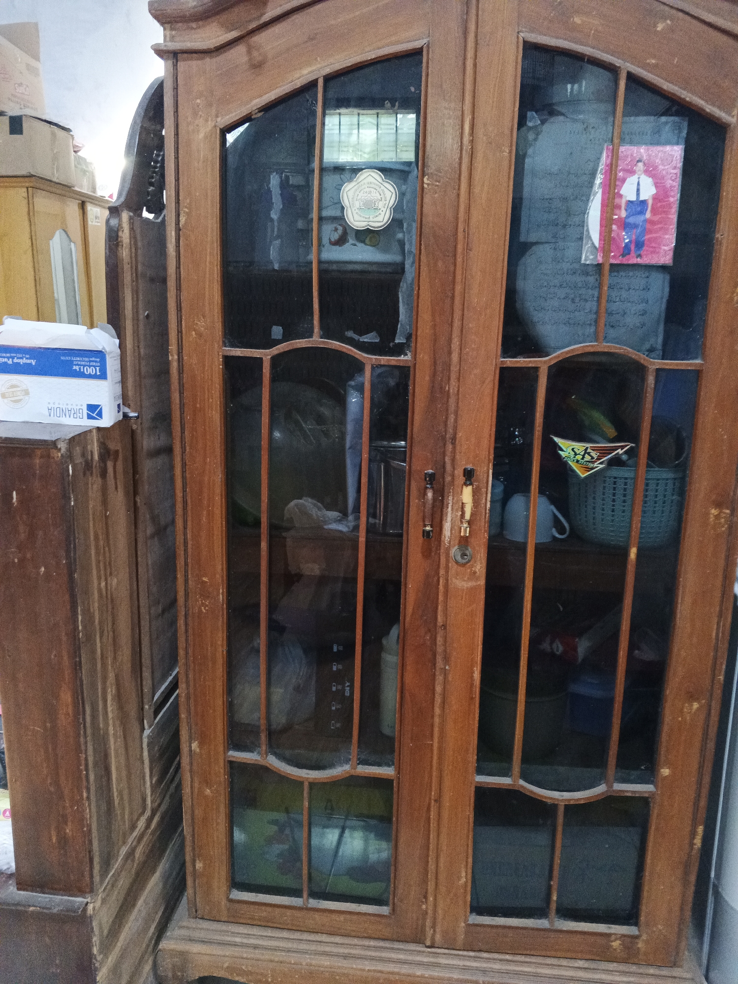 The image shows a closed wooden glass-fronted cabinet in a room in Kemangsen, Indonesia. The cabinet has two doors with arched glass panels, each divided by wooden mullions. Inside the cabinet, several objects are visible, including a stack of books or papers on the left side, and on the right side, a woven basket, a mug, and what appears to be a framed picture of a person. A box labeled "GRANDIA" and "AMPLOP PUTIH" is placed on top of another wooden surface to the left of the cabinet. The environment appears to be dimly lit, with some light filtering in from an unseen source, possibly a window. The overall impression is one of a home interior with storage furniture.