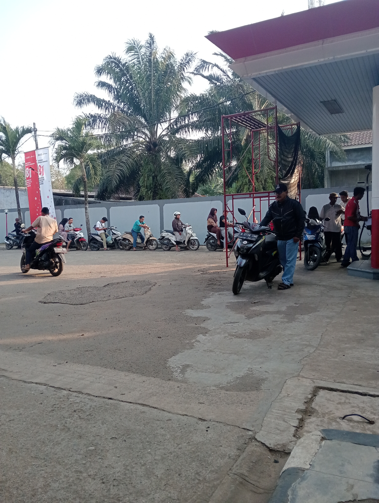 The image captures a scene at a Pertamina gas station in Muara, Indonesia, under a clear sky with palm trees and a red-roofed canopy. Several individuals are present, with the majority riding or standing near motorcycles. In the foreground, a man in a black jacket and blue jeans stands beside a black scooter, looking towards the gas pumps. To his right, another man in a red shirt and blue jeans is actively using a gas pump. Behind them, a line of people on motorcycles are waiting, their vehicles parked parallel to the wall. Among them, a man in a light brown shirt rides a purple scooter. Further to the left, a red and white banner displaying "81" is visible, suggesting a promotional event or anniversary. The ground is paved with concrete, showing some signs of wear and tear, with a large pothole in the middle of the area where the motorcycles are parked. The overall impression is a busy, everyday scene at a local gas station.
