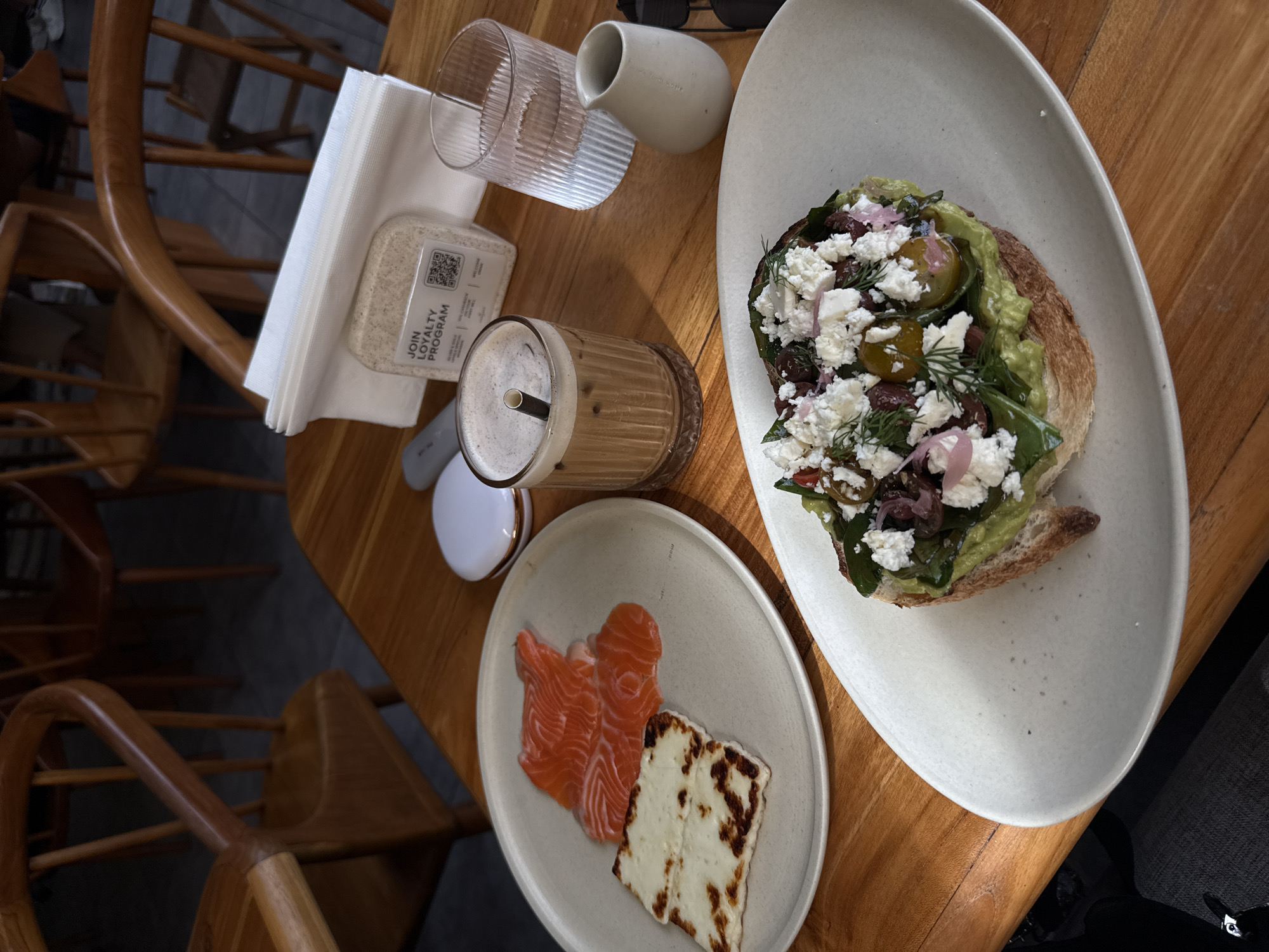 The image shows a wooden table with two plates of food, a glass of water, a small creamer, a drink with a straw, and a stack of napkins. One plate contains avocado toast topped with feta cheese, red onion, and herbs. The other plate contains slices of salmon and grilled halloumi cheese. The scene appears to be inside a restaurant in an unknown location. The table is surrounded by wooden chairs with curved backs.