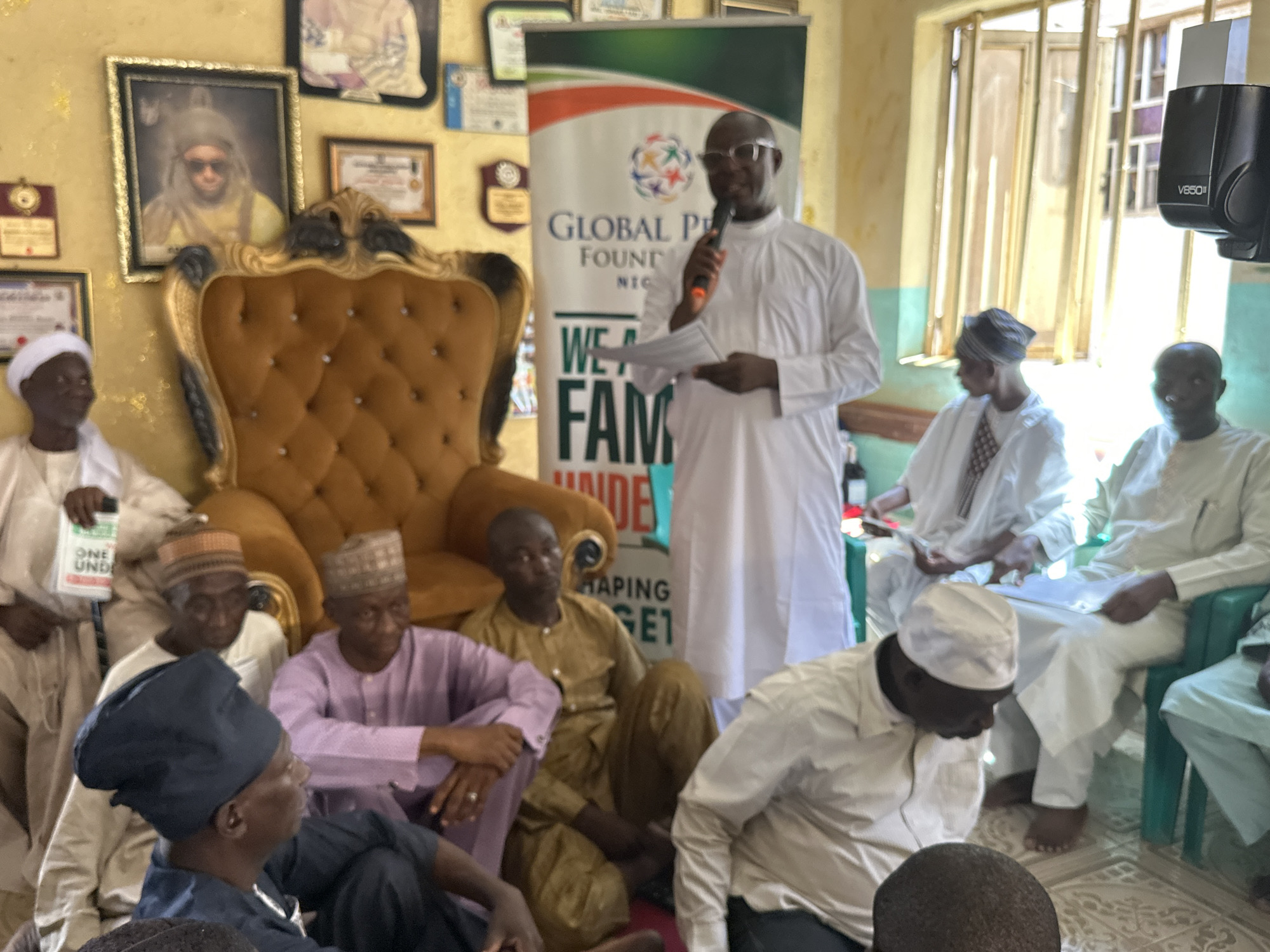 A gathering of men indoors is captured in a medium shot, with the primary focus on an individual in a white robe and glasses speaking into a microphone. He is positioned in front of a banner for the Global Peace Foundation Nigeria. Seated around him on the floor and on chairs are several other men, most of whom are also dressed in white or other light-colored clothing, with various head coverings. Some hold papers and appear to be listening attentively.
Behind the speaker, an ornate gold chair is prominent, and framed portraits and certificates adorn the wall, adding a sense of formality or significance to the setting. Natural light enters through a window to the right of the frame, near which a V650II device is visible. The indoor environment suggests a meeting or presentation of some kind, with the setting located in the city of Unknown, Unknown.