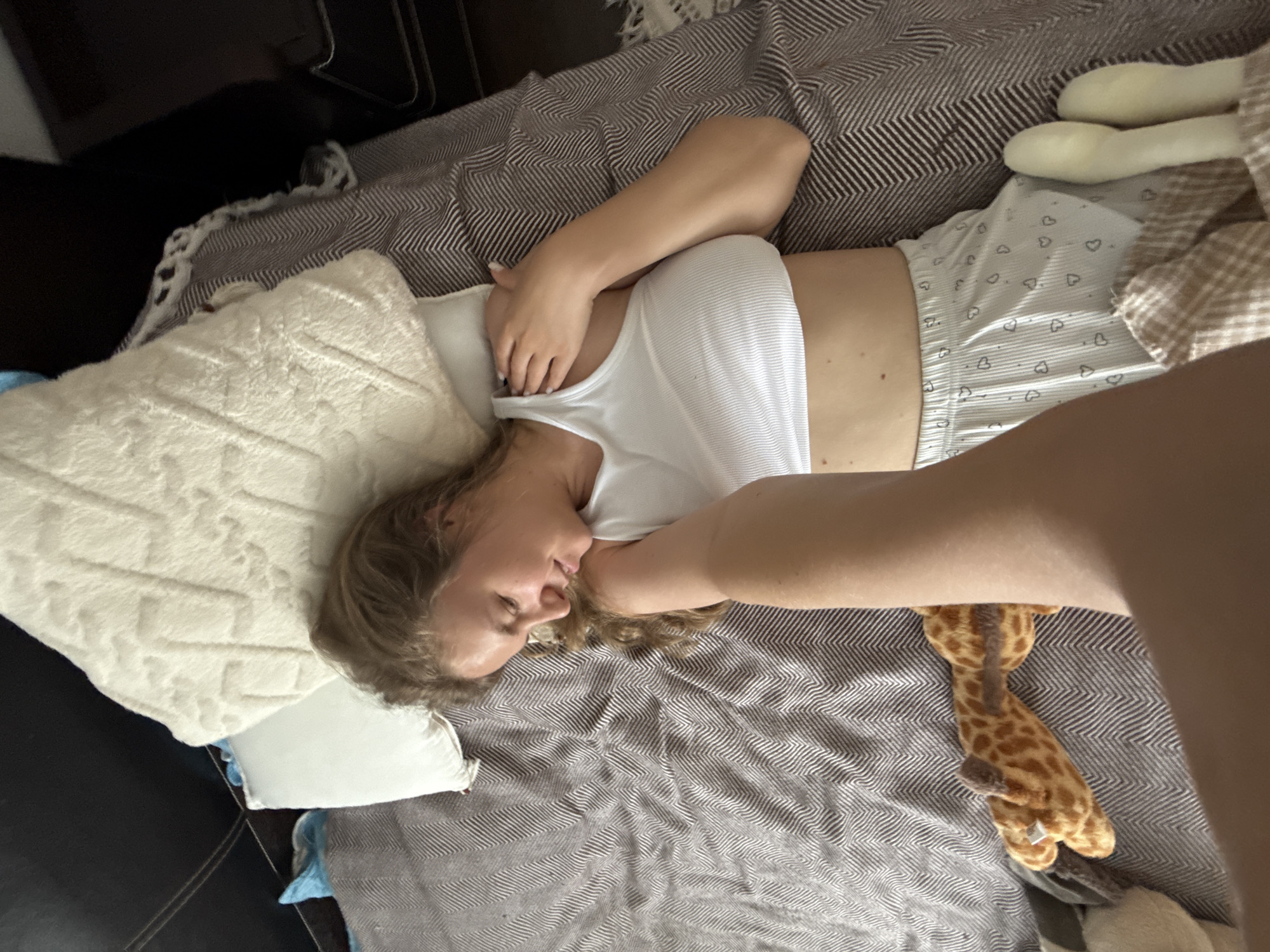 A young woman is lying on her side on a bed. She is wearing a white tank top and grey shorts with a heart pattern. Her arm is bent, and her hand is covering her face. She has blonde, curly hair. The bed is covered with a striped blanket in shades of brown and white. There are several pillows on the bed, including a fluffy white one and a smaller light blue one. A stuffed giraffe is visible near her legs. The lighting suggests it could be daytime. The overall mood appears relaxed or sleepy. The scene is indoors, likely a bedroom. There is no visible text in the image.