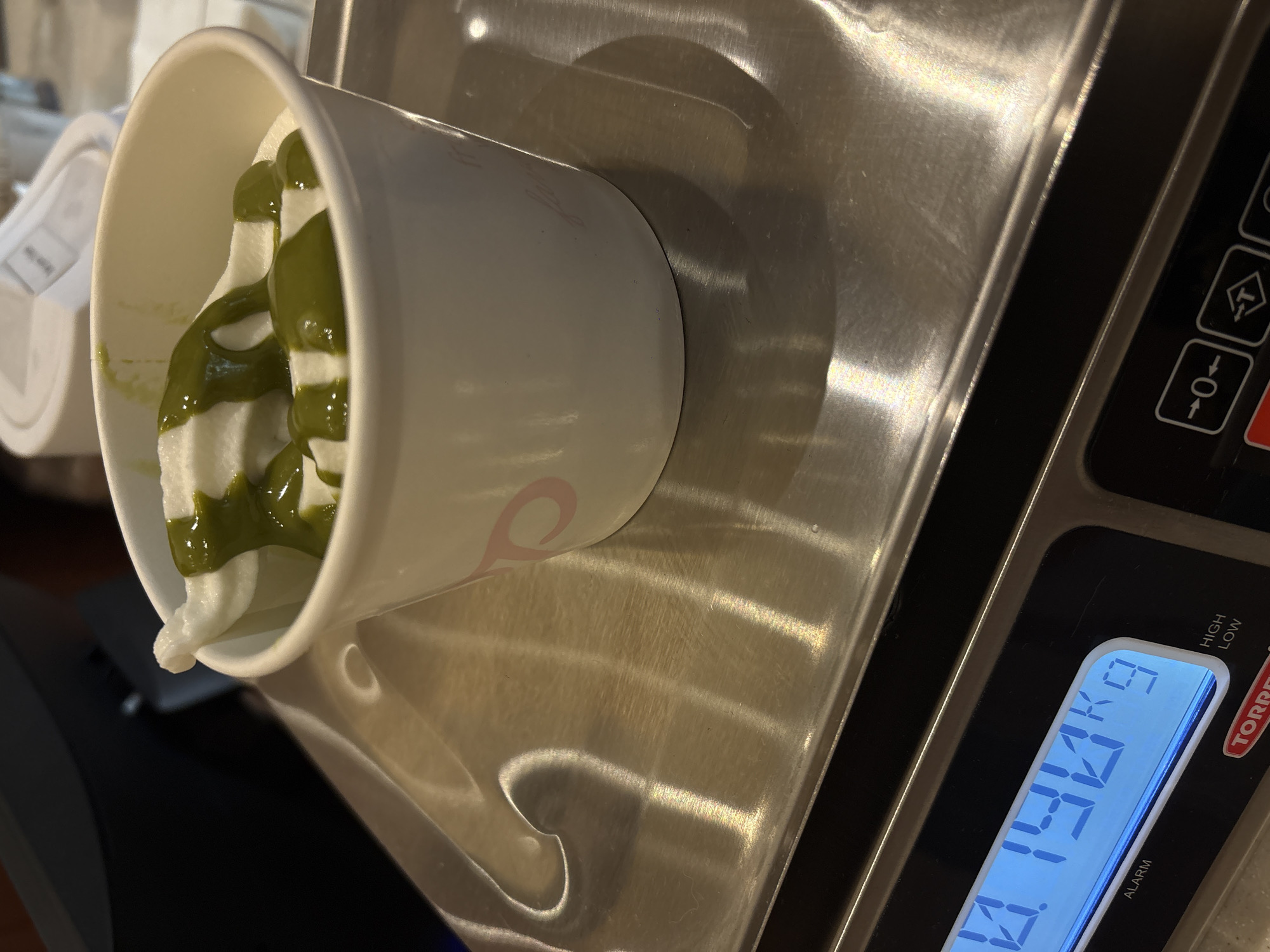 A close-up, high-angle shot captures a paper cup filled with soft-serve ice cream drizzled with green matcha sauce. The cup rests on a stainless steel digital scale, displaying a weight of 0.144 kg. The scale is a Torrey brand, likely used in a commercial or food service setting. The ice cream has a swirled pattern, indicative of being dispensed from a soft-serve machine. Based on the presentation and content, this scene is likely taking place in a dessert shop in Mexico City, Mexico.
