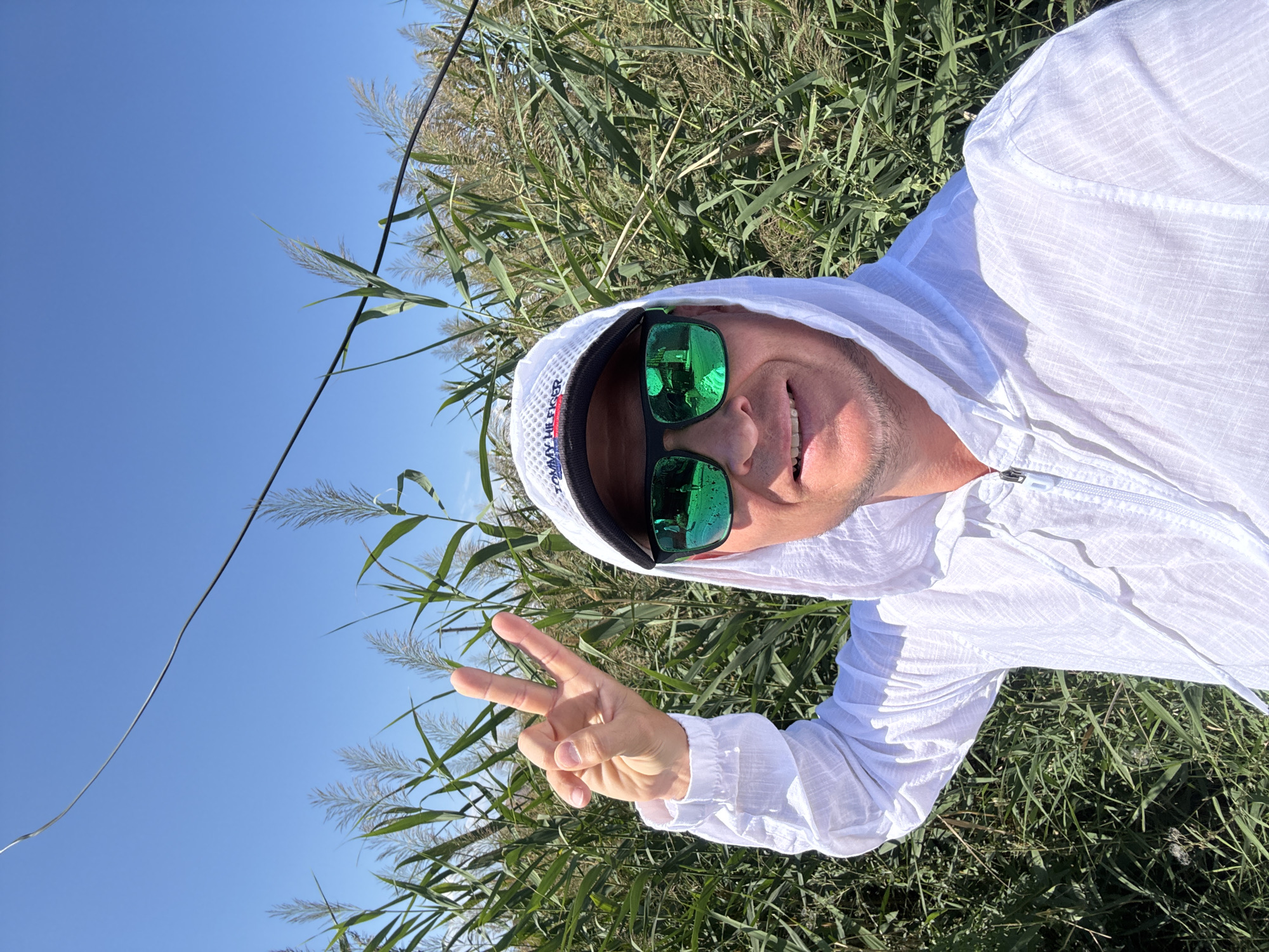 A man is taking a selfie outdoors in Zharbulak, Kazakhstan. He is wearing a white hooded jacket, sunglasses, and a white baseball cap with the Tommy Hilfiger logo. He is smiling and holding up two fingers in a peace sign. Behind him, there is a field of tall green reeds under a clear blue sky. A dark wire is visible in the upper portion of the image.