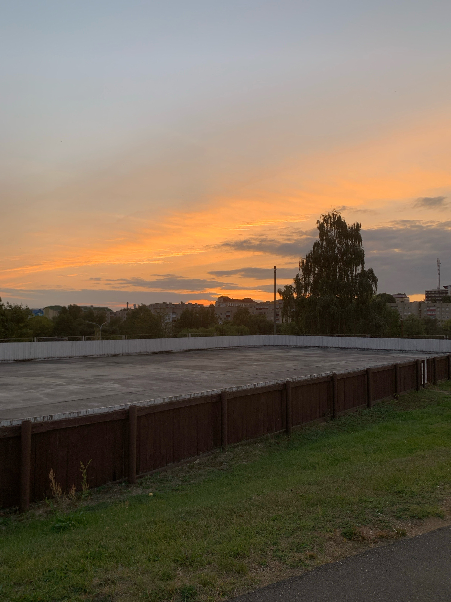 The image shows a wide shot of an outdoor ice rink or court, enclosed by a wooden fence. The surface of the rink appears to be concrete, with some wear and tear visible. A wooden fence, made of vertical planks, runs along the perimeter of the rink. To the right of the rink, there is a grassy area with some weeds. In the background, there is a line of trees and residential buildings, characteristic of a city. A tall, slender tree with drooping branches stands out in the middle ground, silhouetted against the sky. The sky is filled with a sunset, displaying vibrant orange and yellow hues, with some scattered clouds. The overall atmosphere is serene and peaceful, with the warm tones of the sunset creating a calming effect. The location is identified as the city of Kapyĺ, Belarus. There are no people or animals visible in the image. The lighting suggests it is late afternoon or early evening.