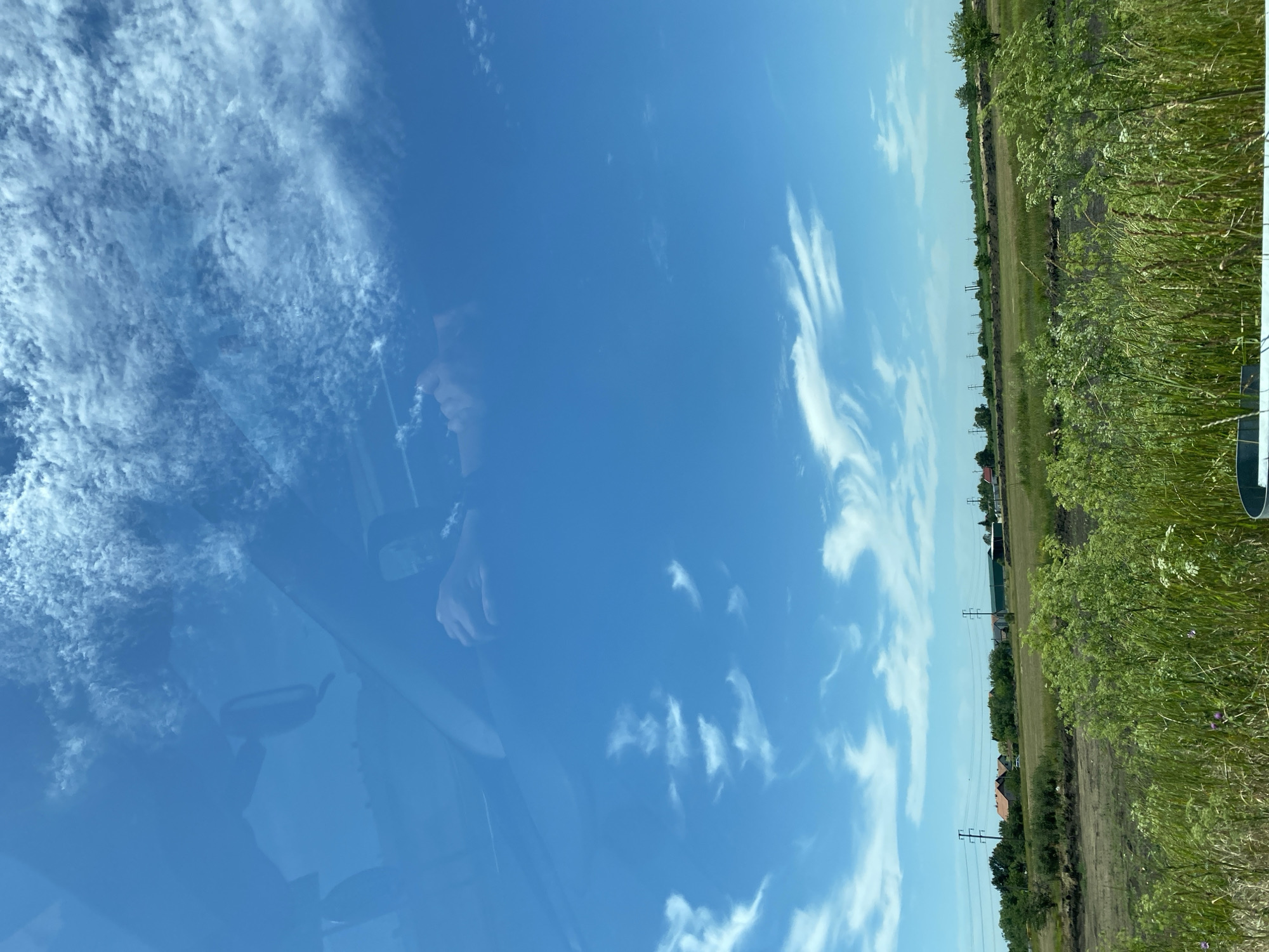 The image shows a view from inside a vehicle looking out through the windshield. The sky is a vibrant blue with scattered wispy white clouds. In the reflection on the windshield, parts of the car's interior are visible, including a rear-view mirror and a person's hands on the steering wheel.
Beyond the windshield, a rural landscape unfolds. A grassy field stretches towards a row of houses with red-tiled roofs and some trees. Power lines with tall poles are also visible in the middle ground. The foreground consists of lush green vegetation, including tall grasses and some flowering plants.
The scene appears to be in a suburban or rural area, possibly on the outskirts of Zrenjanin, Serbia, given the context provided. The lighting suggests it is daytime, likely under clear or partly cloudy skies. There are no overt signs of activity or specific events occurring in the image, beyond the implied presence of the vehicle and its occupants.
