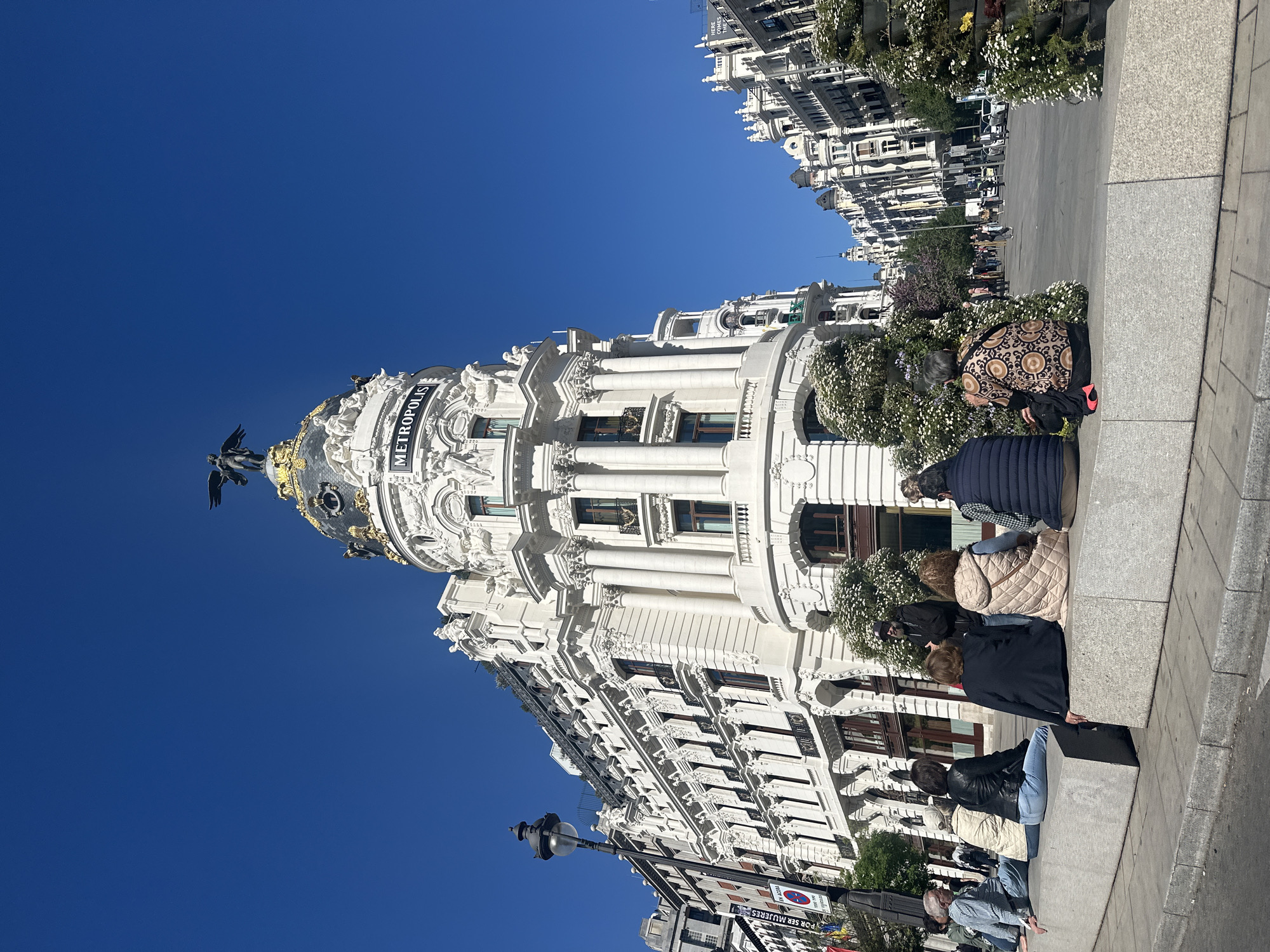 The image shows the Metropolis Building in Madrid, Spain, with a clear blue sky in the background. The building is predominantly white and features ornate architectural details. A group of people are sitting on a low wall adjacent to the building, facing away from the camera. Flowers are planted between the building and the wall. A street with other buildings is visible in the distance. A street lamp and some street signs are also visible.