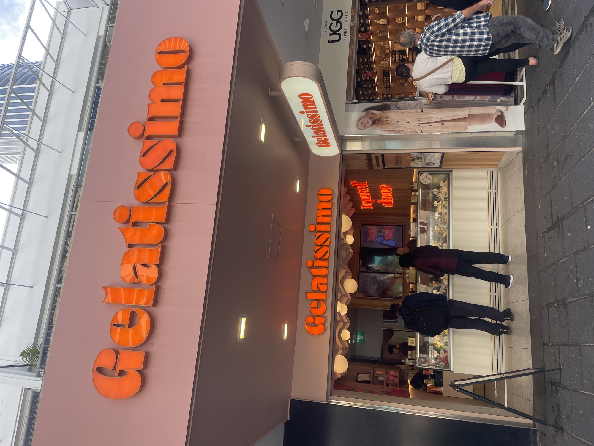 In Surfers Paradise, Australia, a storefront for "Gelatissimo" is prominently displayed. The name "Gelatissimo" is emblazoned in large, vibrant orange, three-dimensional lettering across a pale pink facade above the entrance. Beneath this, a smaller, illuminated oval sign also displays "Gelatissimo" in orange lettering. The shop appears to be a gelato parlor with a display case showcasing various flavors of gelato. Inside, staff members are visible behind the counter. Customers are seen in front of the counter, some browsing and making selections. To the right of the gelato shop, there is a neighboring store with a sign for "UGG" and displays of footwear. The street in front of the shops is paved with dark grey cobblestones. Two individuals, one wearing a blue and white checkered shirt and another in a white top and black pants, are walking away from the viewer towards the right side of the frame, appearing to be customers. A third person, mostly obscured, is walking ahead of them. A large promotional display featuring a woman in a fuzzy robe stands near the entrance of the gelato shop. The overall environment suggests a commercial street scene in a popular tourist area.