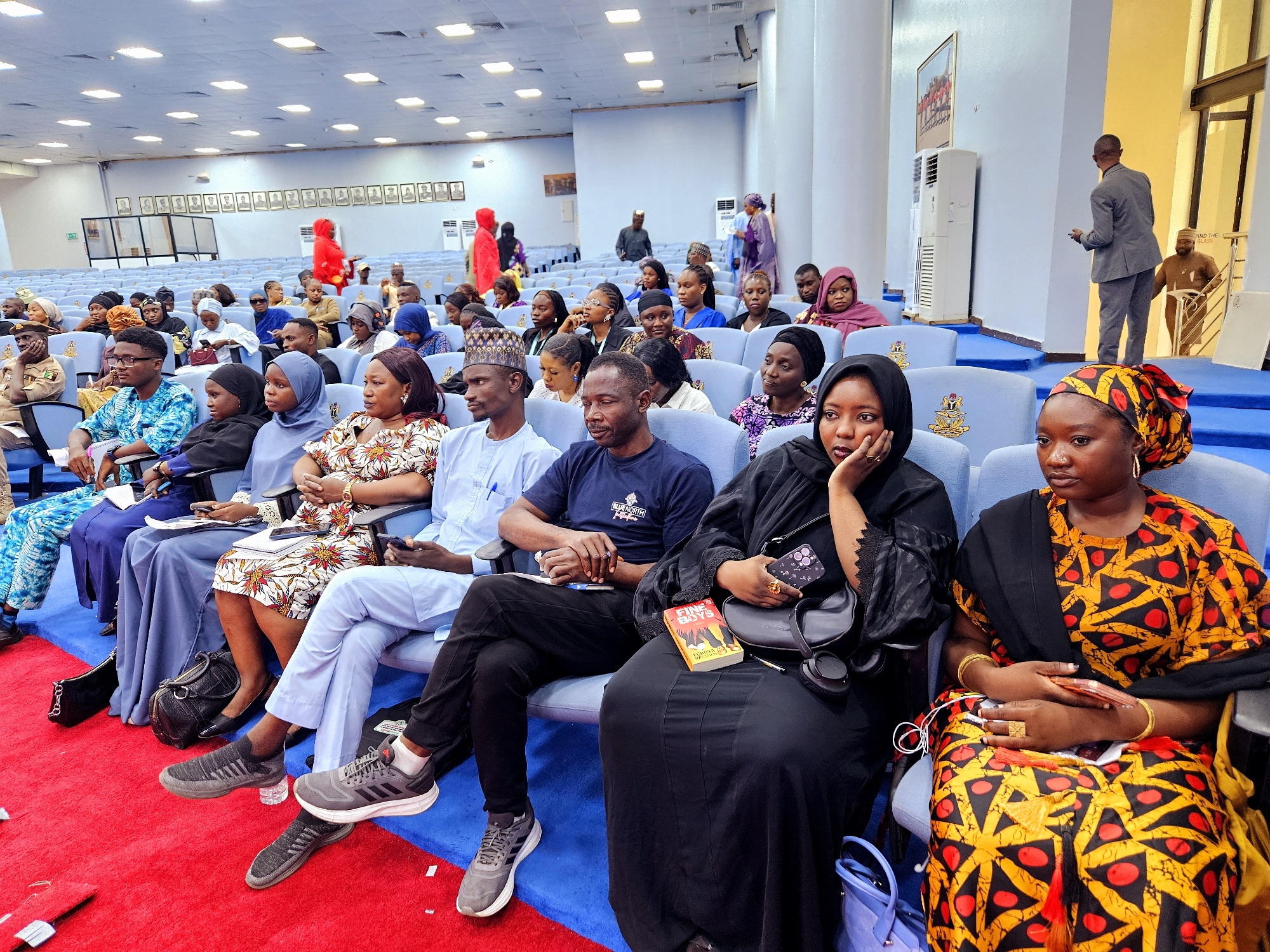 In an indoor auditorium in Gwarinpa, Nigeria, approximately thirty individuals, a mix of men and women, are seated in rows of blue chairs on a blue carpeted floor. The seated audience appears to be attending an event, with some individuals looking towards an unseen focal point. A red carpet runs diagonally across the foreground.
In the front row, from left to right:
A woman in a black hijab and dress rests her chin on her hand, looking to her left. Next to her, a woman in a vibrant yellow and black patterned dress, adorned with red circles, sits with her hands clasped in her lap. She is wearing gold jewelry, including bangles and rings. A book titled "FINE BOYS" is visible on the lap of the woman next to her.
In the second row, from left to right:
A man in a dark blue t-shirt and black pants sits with his hands clasped. A woman in a black hijab and dress is seated next to him, holding a phone. A woman in a light blue hijab and dress is also in this row.
Further back, the audience continues, with many individuals wearing headscarves. In the background, a man in a grey suit is visible near a staircase, gesturing with his right hand. Two large air conditioning units are mounted on the wall. Several framed pictures hang on the wall above the audience. The lighting in the room is bright, coming from overhead fluorescent lights. The overall scene suggests an organized gathering or conference.