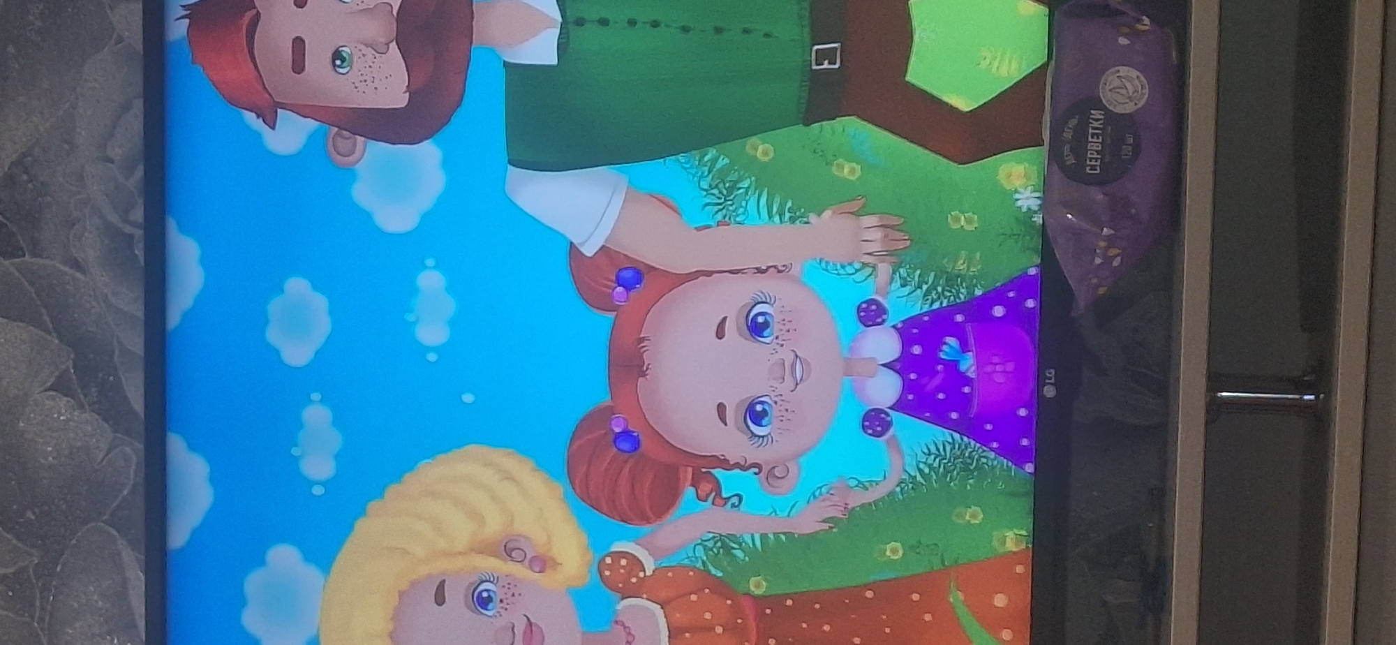 An animated family, consisting of a man, a woman, and a young child, are depicted against a bright blue sky with scattered white clouds. The man, on the right, has a red beard and is wearing a green vest over a white shirt. The woman, on the left, has blonde curly hair and is wearing an orange polka-dot dress. The child, in the center, has red hair styled in two buns and is wearing a purple polka-dot dress with a white collar. The child is holding the hands of both the man and the woman. They are standing in a grassy area with some yellow flowers. In the foreground, a dark television screen displays the animated scene, and below the screen is a shelf with a purple package of wet wipes labeled in Russian. The scene is set in Bila Tserkva, Ukraine.