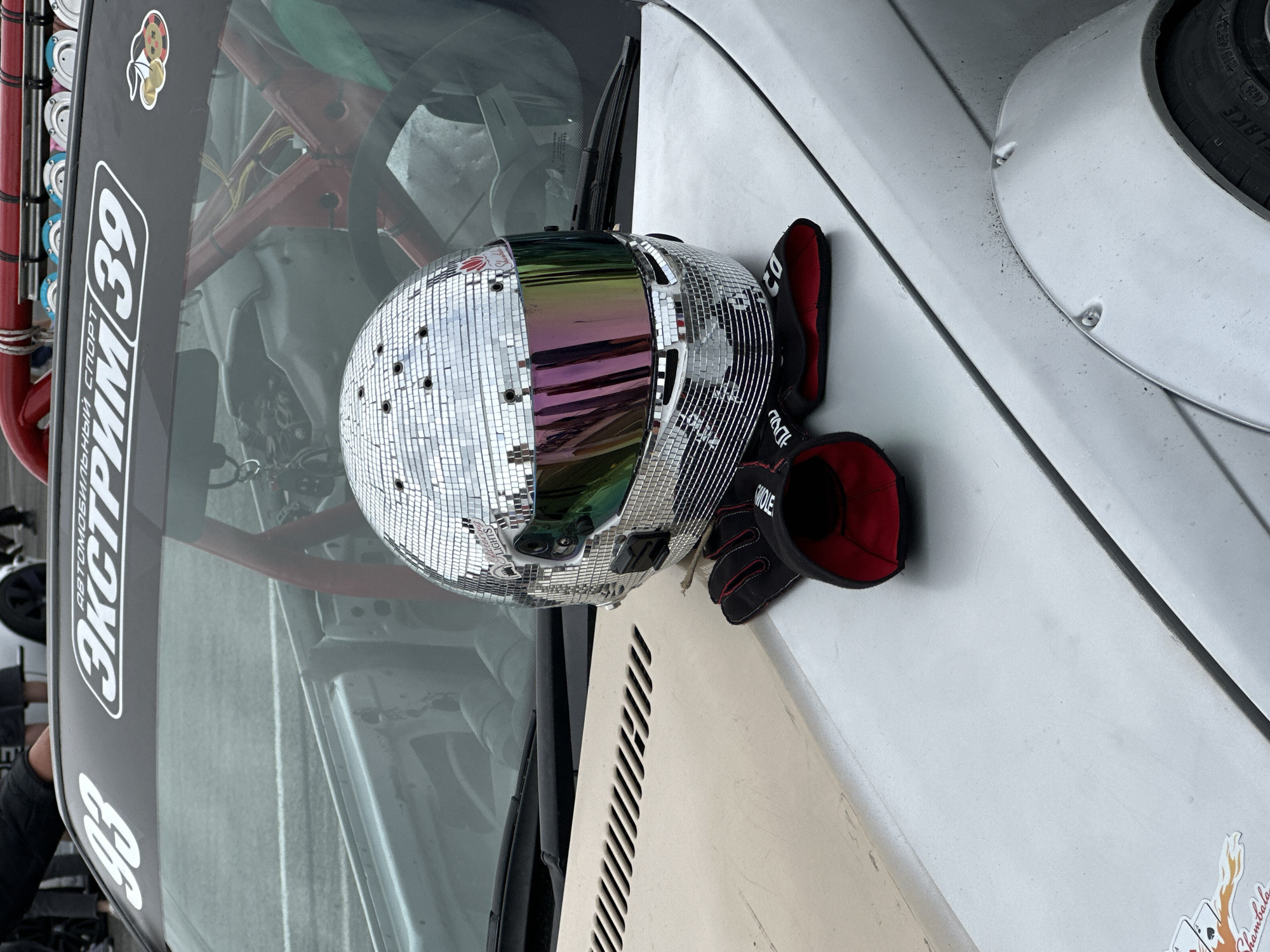 A disco ball-style helmet and a pair of racing gloves are placed on the hood of a silver car. The car has a roll cage visible inside. A sticker on the windshield reads "93 ЭКСТРИМ 39". The scene appears to be outdoors, possibly at a racing event. The presence of the sticker "39" suggests the location is Kaliningrad Oblast, Russia, as the region code is 39.