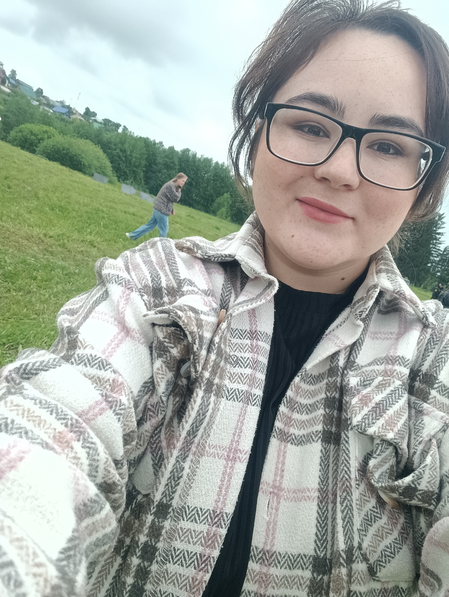The image is a selfie taken outdoors, featuring a young adult female in the foreground. She has dark hair, dark-framed glasses, and freckles, and is wearing a black ribbed top under a multi-colored plaid overshirt with white, pink, and grey tones in a herringbone weave. She looks directly at the camera with a slight smile. In the mid-ground, a second individual, appearing to be female with light hair, is running or walking across a green grassy slope, dressed in a dark patterned top and blue jeans. The background consists of a large green hillside dotted with trees and bushes, with a distant cluster of houses or buildings visible on another elevated area under an overcast sky. The overall scene suggests a rural or semi-rural environment.