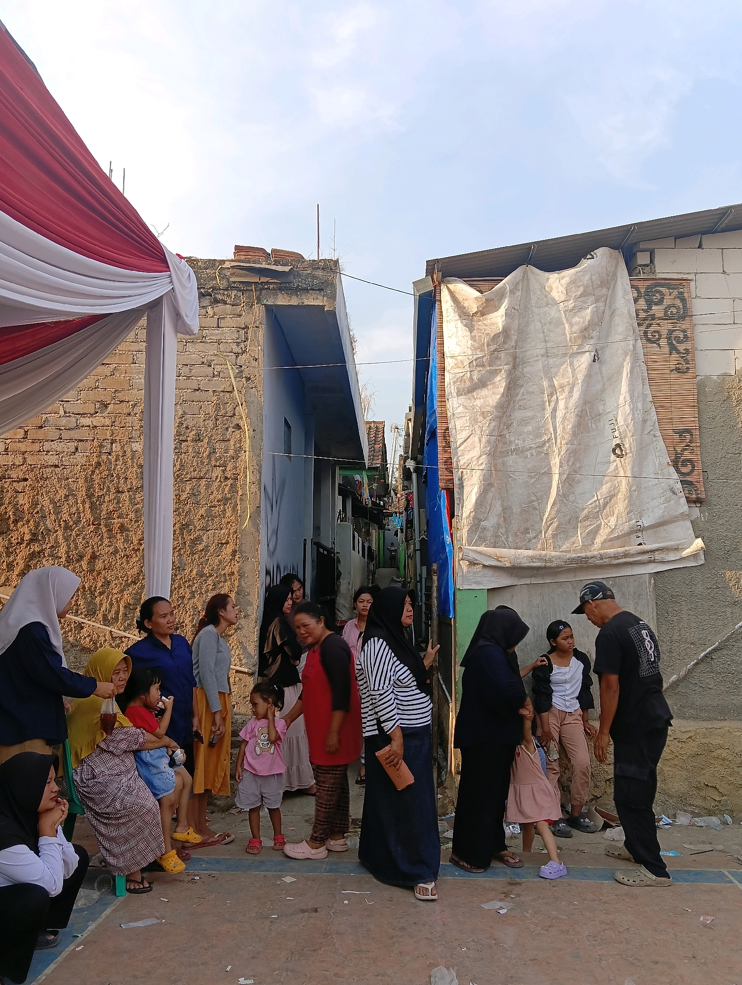 In Padalarang, Indonesia, a group of people gather outdoors. A red and white tent adorns the left side of the frame. To the right, two buildings stand side-by-side. The building on the left is constructed of rough brick, while the building on the right is made of lighter-colored blocks, partially covered by a white tarp and a bamboo mat. The ground is a paved surface. Several individuals are present, including women and children. Some are standing, while others are seated or crouching. The scene appears to be a casual gathering, possibly a community event.