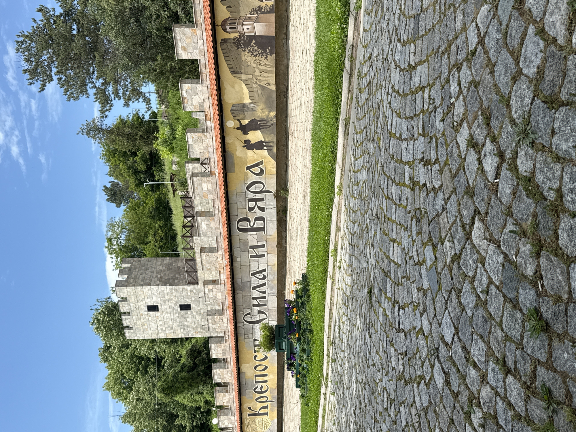 The image depicts a section of the Baba Vida fortress in Vidin, Bulgaria. The shot includes a stone paved walkway, a section of the fortress wall with battlements, and a mural. The mural depicts figures on horseback. The text "Крепост Баба Вида" is displayed vertically along the wall. Trees and vegetation are visible behind the fortress wall. The sky is blue.