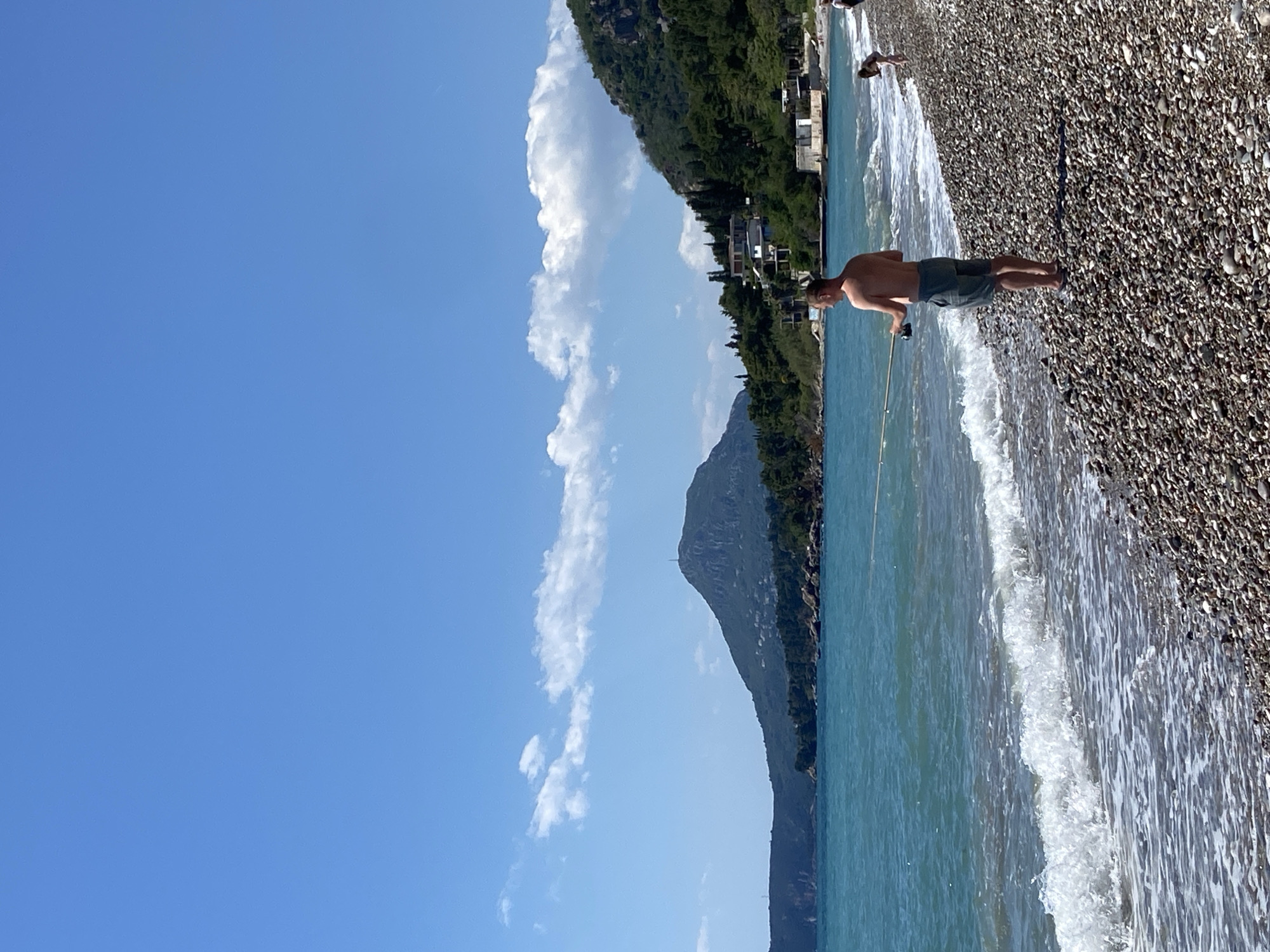 A man stands on a pebble beach by the sea, holding a fishing rod. The sea is a light turquoise color, with small waves breaking on the shore. Behind the sea is a steep, green mountain with houses dotted along its slopes. The sky is blue with some white clouds. The location is likely near the city of Bar, Montenegro.