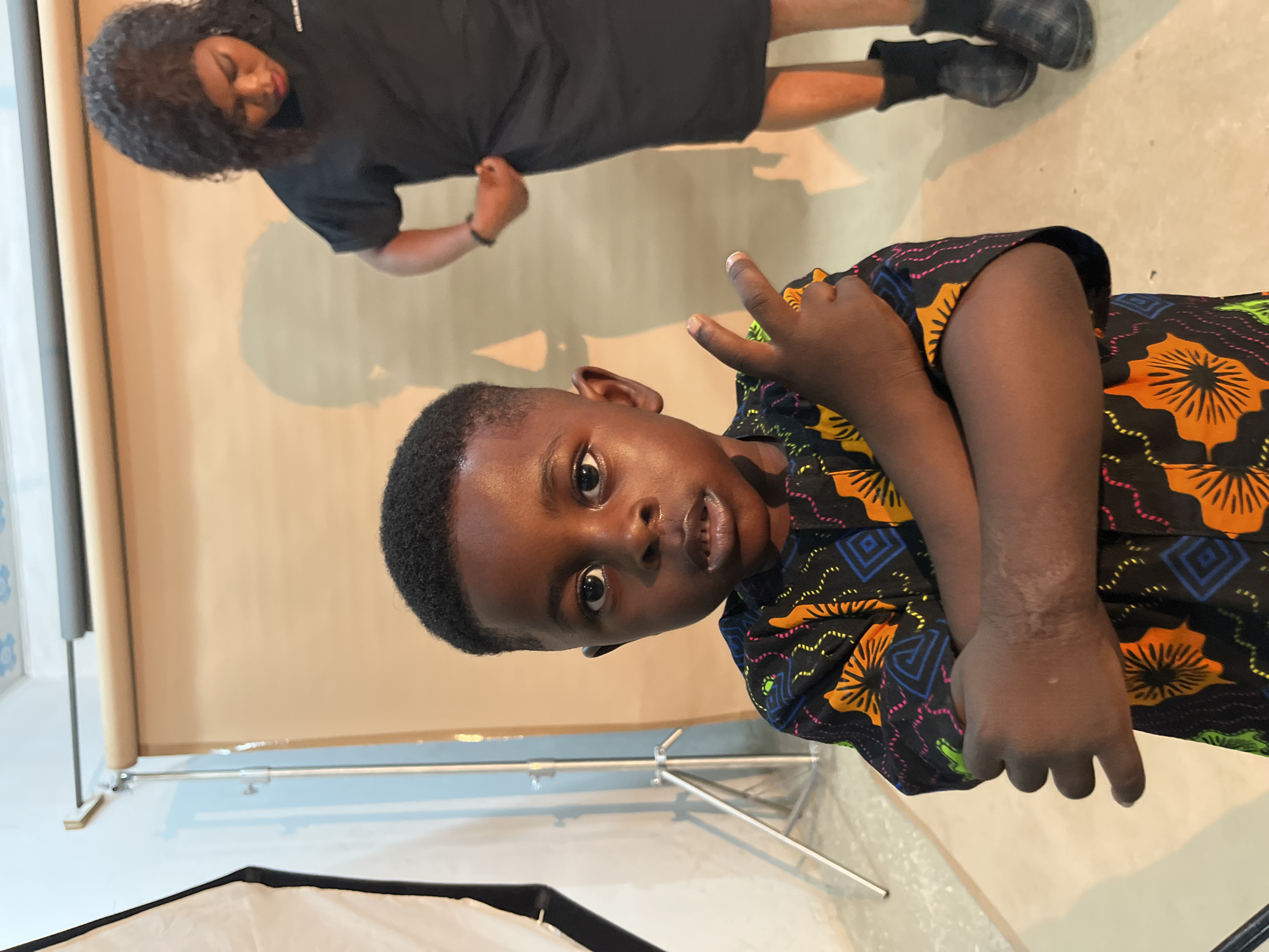The media file shows two people in what appears to be a photo studio. In the foreground, a young boy is looking directly at the camera with a surprised expression. He has short black hair and is wearing a colorful dashiki with orange, yellow, blue, and pink geometric patterns and floral motifs. His right hand is raised, and he is making a "rock on" gesture with his index and pinky fingers extended. His left arm is resting on his lap.
In the background, a person with long, curly black hair is wearing a black t-shirt and black shorts. Their head is tilted down, and they appear to be looking at their feet, which are clad in black shoes. This person is standing, and their shadow is visible on the light brown backdrop behind them.
The setting includes a light brown backdrop on a stand, and a white photography softbox is visible in the lower left corner. The floor is a light concrete. The lighting suggests an indoor environment, likely during the daytime given the brightness. The overall atmosphere appears to be casual and playful, especially with the boy's expression and gesture. No visible text is present in the image. The location context is Gura Top, Nigeria.
