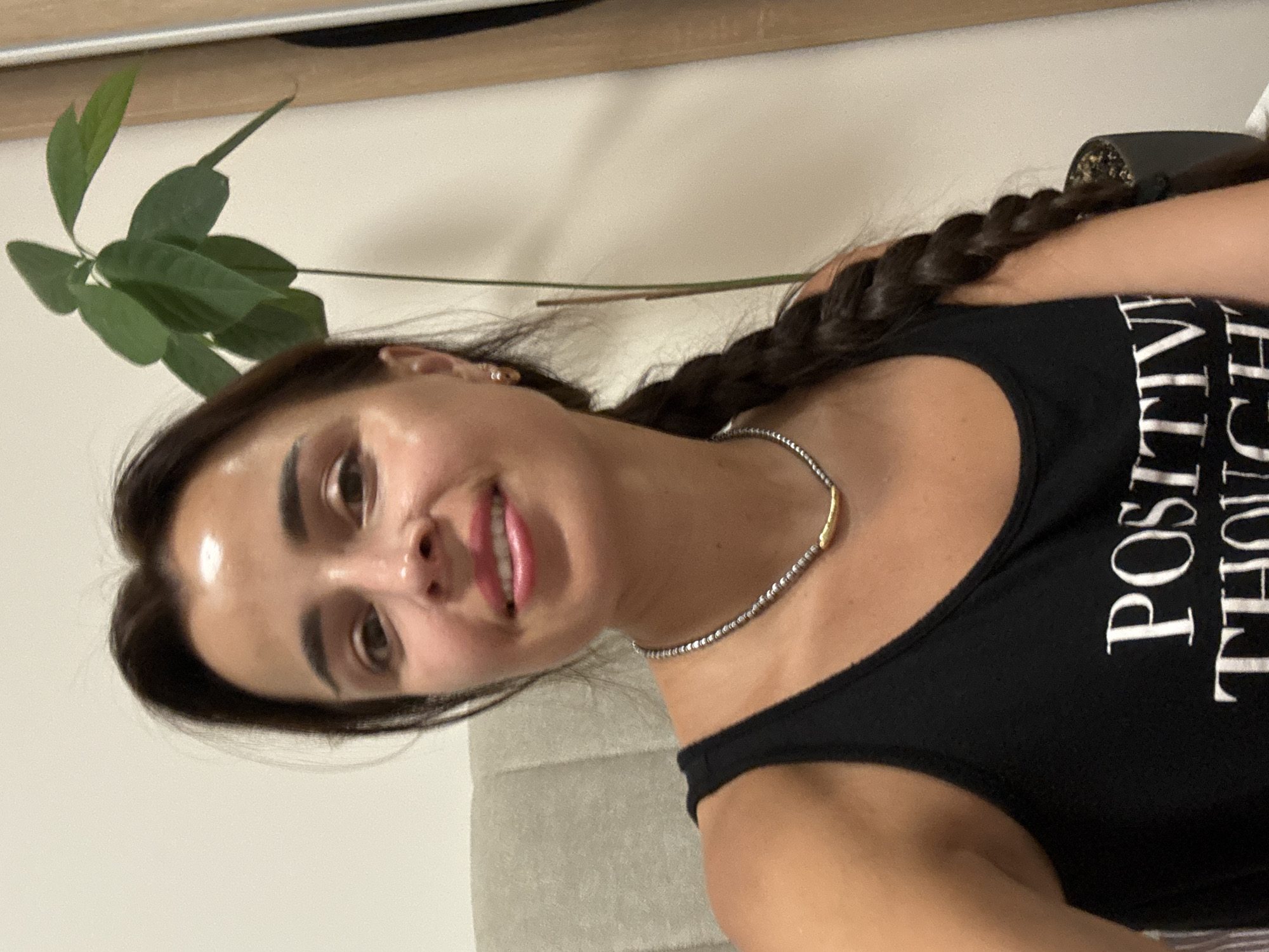 A woman with dark hair in a braid, wearing a black tank top and a necklace, smiles at the camera. She is seated in front of a chair, with a plant and a wall behind her. The image is a medium shot, taken from the chest up.