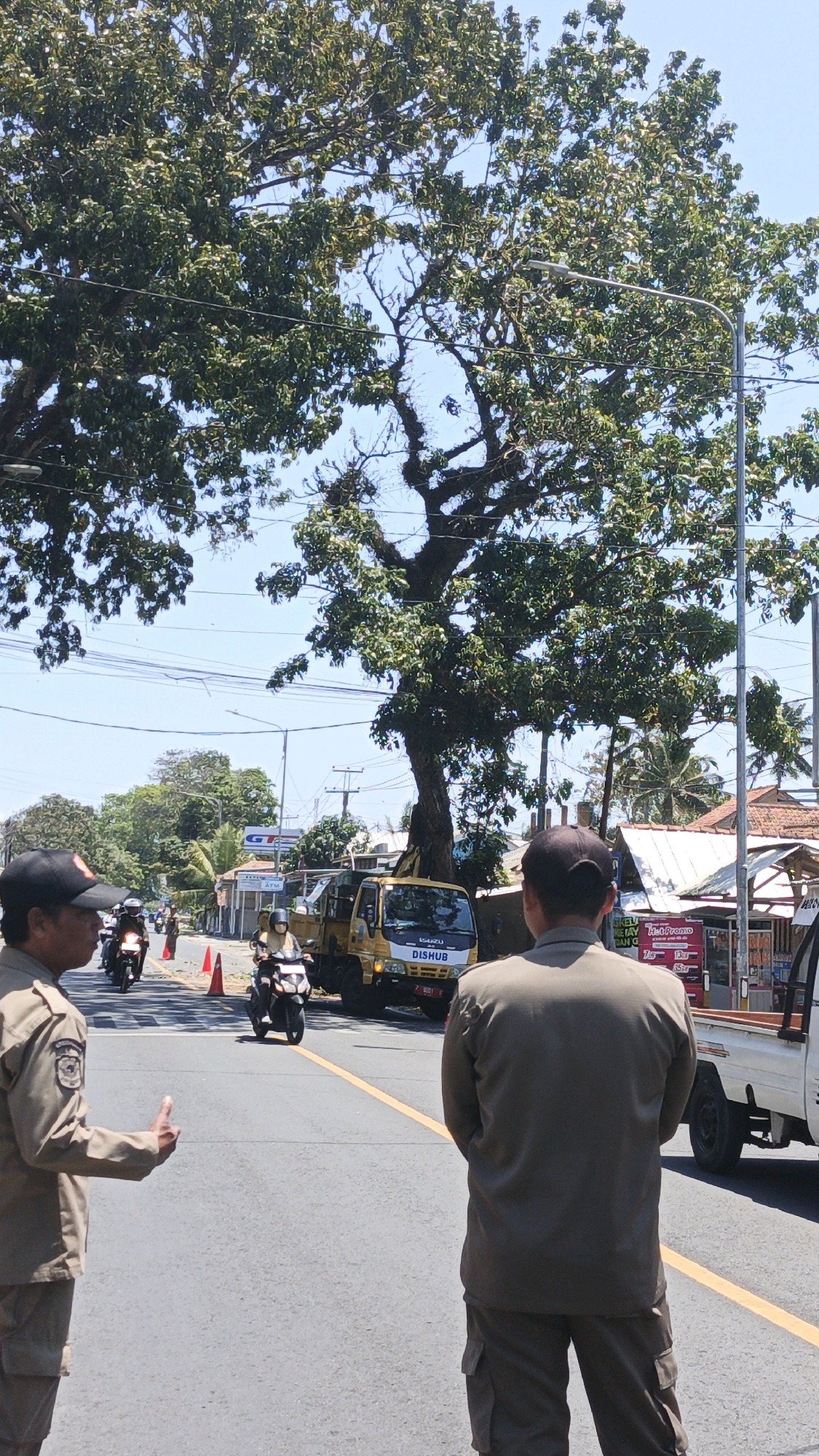 Two uniformed men, likely traffic officers, stand on a paved road. The man on the left, facing the camera, wears a tan uniform, a black cap, and a badge on his left sleeve. He gestures with his right hand, palm open, as if directing traffic. The man on the right faces away from the camera, wearing a similar tan uniform. In the background, a yellow truck with "DISHUB" written on its side is stopped on the road. Several motorcycles are also visible, some with riders. A large tree with dense green foliage dominates the upper portion of the image. The scene appears to be set in a city environment with buildings and power lines present. The overall atmosphere is bright and sunny, suggesting it is daytime. The location is specified as Sukaresik, Indonesia.