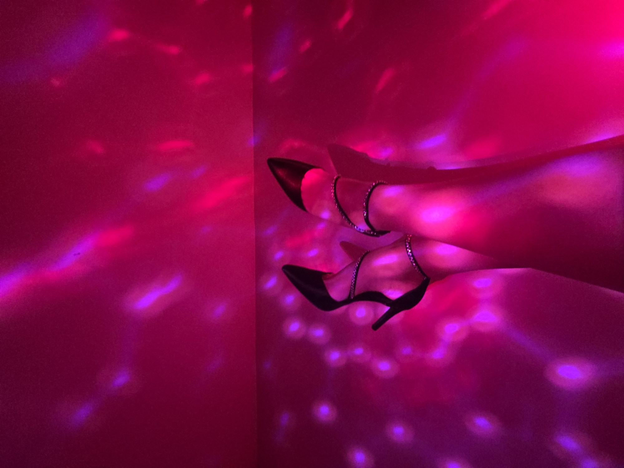 A close-up, indoor shot depicts two legs wearing black high heels with silver ankle straps, positioned in front of a corner. The legs are illuminated with a bright pink and purple light, creating a disco-like ambiance. The location is Unknown, Unknown.