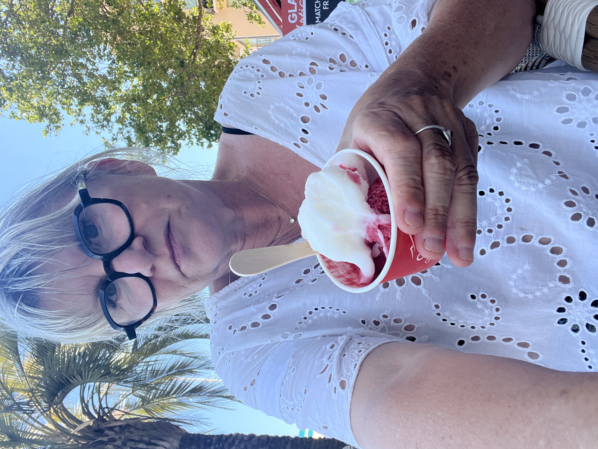 A close-up, low-angle, portrait orientation shot shows a woman holding a red paper cup filled with red and white sorbet or ice cream. She is wearing a white eyelet top and has short, blonde hair and black-rimmed glasses. Her right hand, adorned with a ring on her index finger, holds the cup, and a small wooden spoon is partially submerged in the sorbet. The background reveals a bright blue sky, green foliage from trees, and the suggestion of buildings, possibly in an urban setting. The sunlight suggests it's daytime, likely warm weather. A palm tree frond is visible in the lower left corner. Text is visible on a red sign in the upper background, reading "GLAM MATCH FR...". The overall impression is a candid moment of someone enjoying a treat outdoors on a sunny day in Cannes, France.