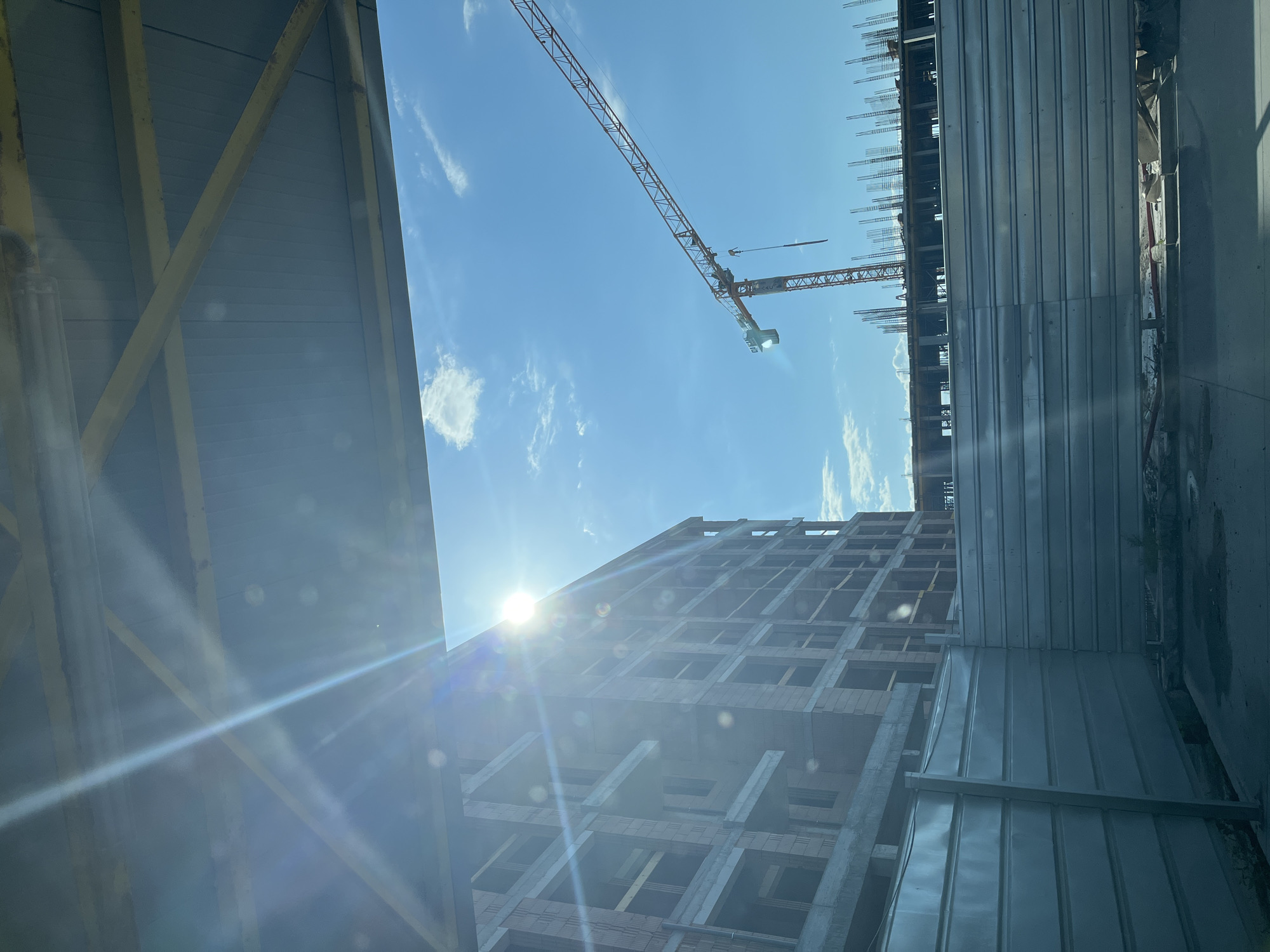 A construction site in Plovdiv, Bulgaria, is captured on a clear, sunny day. The image is framed at the top by the underside of a light blue-grey corrugated metal canopy or roof structure, supported by yellow-painted metal beams, with prominent sun flare radiating from the upper left quadrant. To the left, a multi-story building is under construction, featuring exposed reddish-brown ceramic blocks forming walls and grey concrete slabs for floors and balconies. The sun's glare partially obscures the top of this building. To the right, an orange tower crane, with "CAEL" visible on its mast, extends its long arm diagonally across the bright blue sky, which contains scattered white clouds. Below the crane, another section of the construction site shows concrete foundations or floor slabs with numerous vertical steel rebars protruding upwards. A light grey, horizontally corrugated metal fence acts as a perimeter barrier in the lower portion of the image, separating the construction area from the immediate foreground, which shows a concrete or dirt surface. No individuals are visible in the scene.