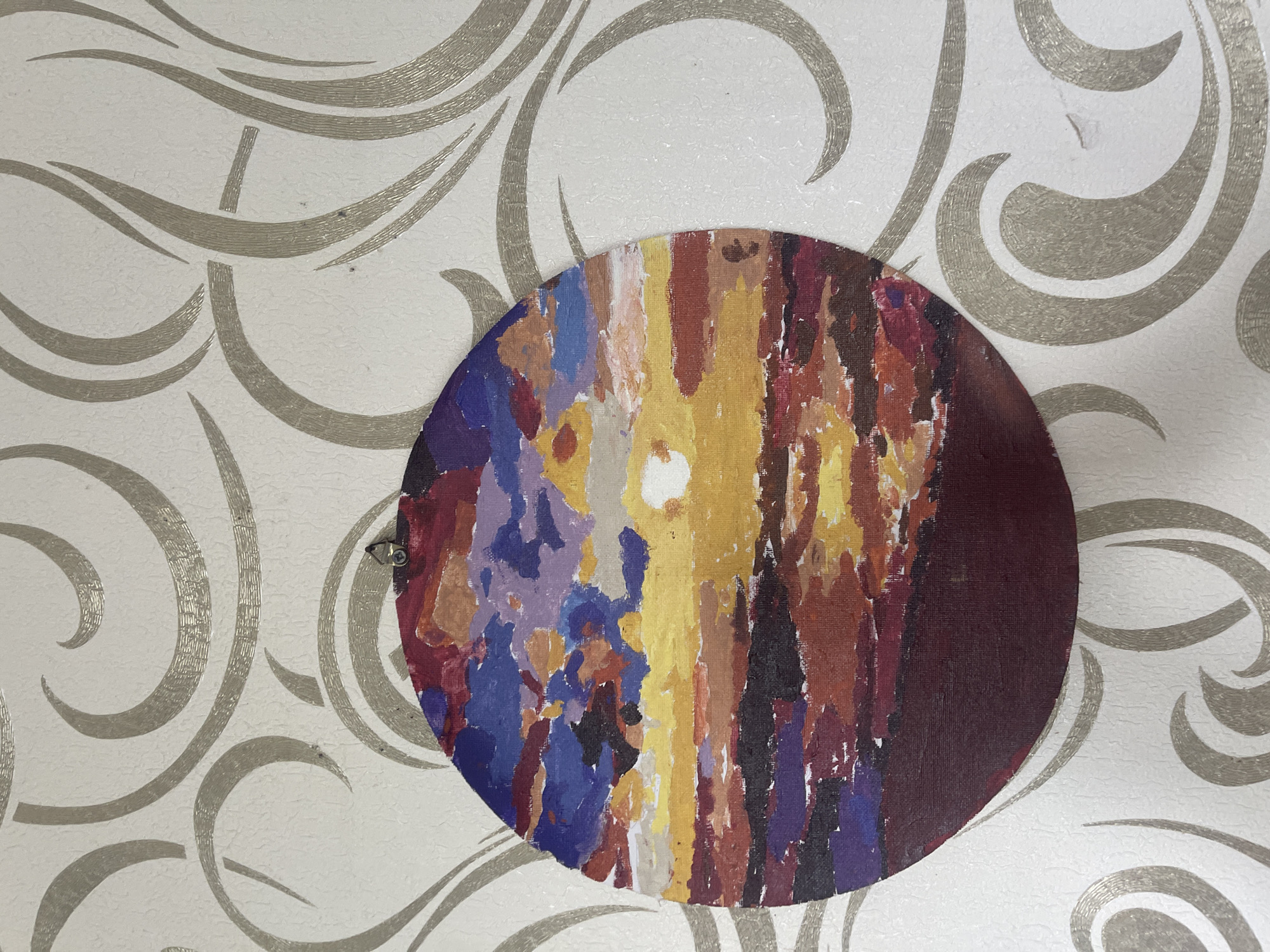 An abstract circular painting of a sunset is displayed on a wall with a textured wallpaper. The painting features bold brushstrokes and a vibrant color palette, with shades of yellow, orange, red, purple, and blue. The sunset is depicted with a bright yellow sun at the center, radiating outwards with fiery colors. The wallpaper has a subtle damask pattern in a light, almost white, color with a slight shimmer. The painting is hung by a single screw at the top.